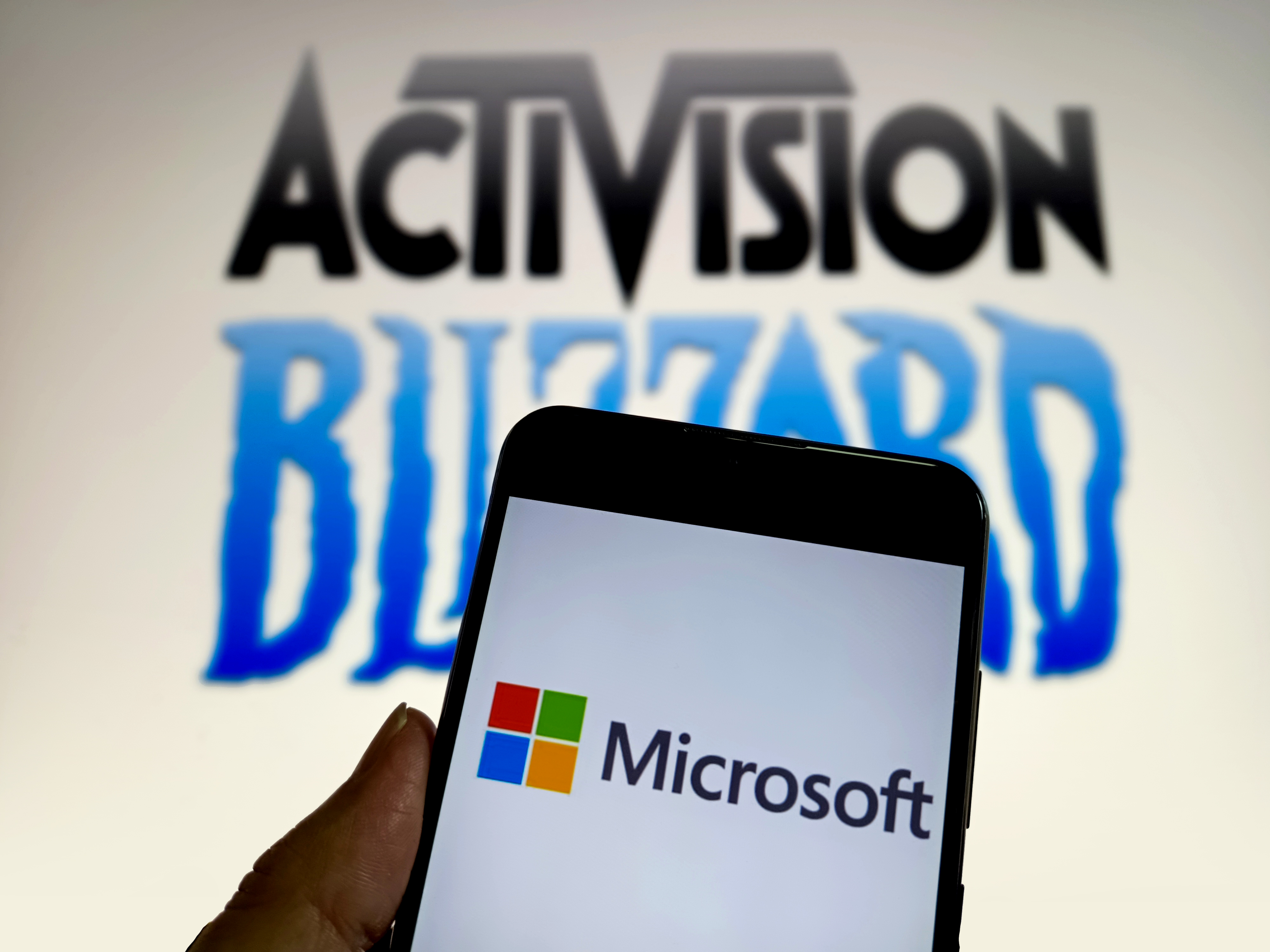 A phone with the Microsoft logo is held up in front of the Activision Blizzard logo, illustrating Microsoft’s acquisition deal.