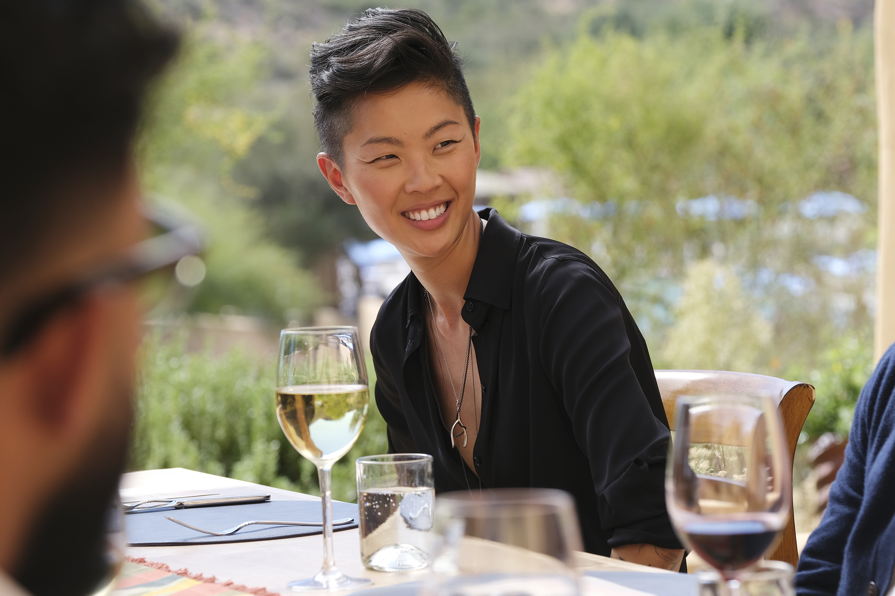 Kristen Kish smiles in front of a background of faraway green trees during an episode of Top Chef Season 19. She wears a black shirt. There is a full glass of white wine in front of her. 