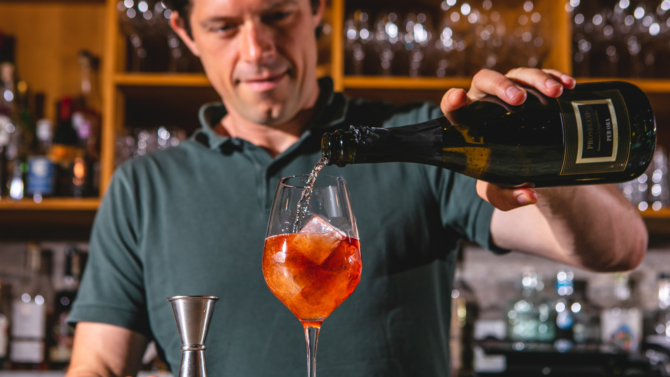 A man at a bar pours a bottle into a glass with a negroni-colored drink in it. 