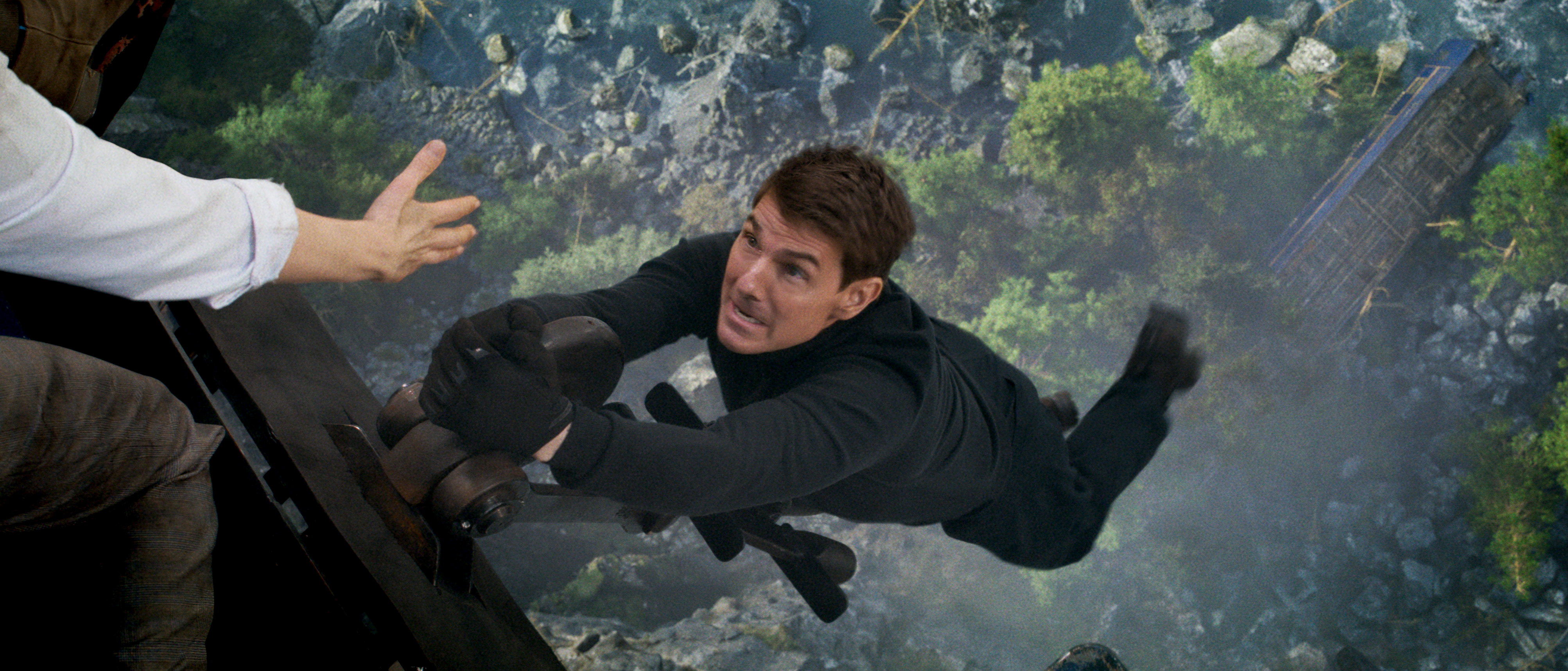 Tom Cruise as Ethan Hunt hanging off the edge of a train with a ravine very far below him as an outstretched hand from someone offscreen reaches for him in Mission: Impossible — Dead Reckoning Part 1