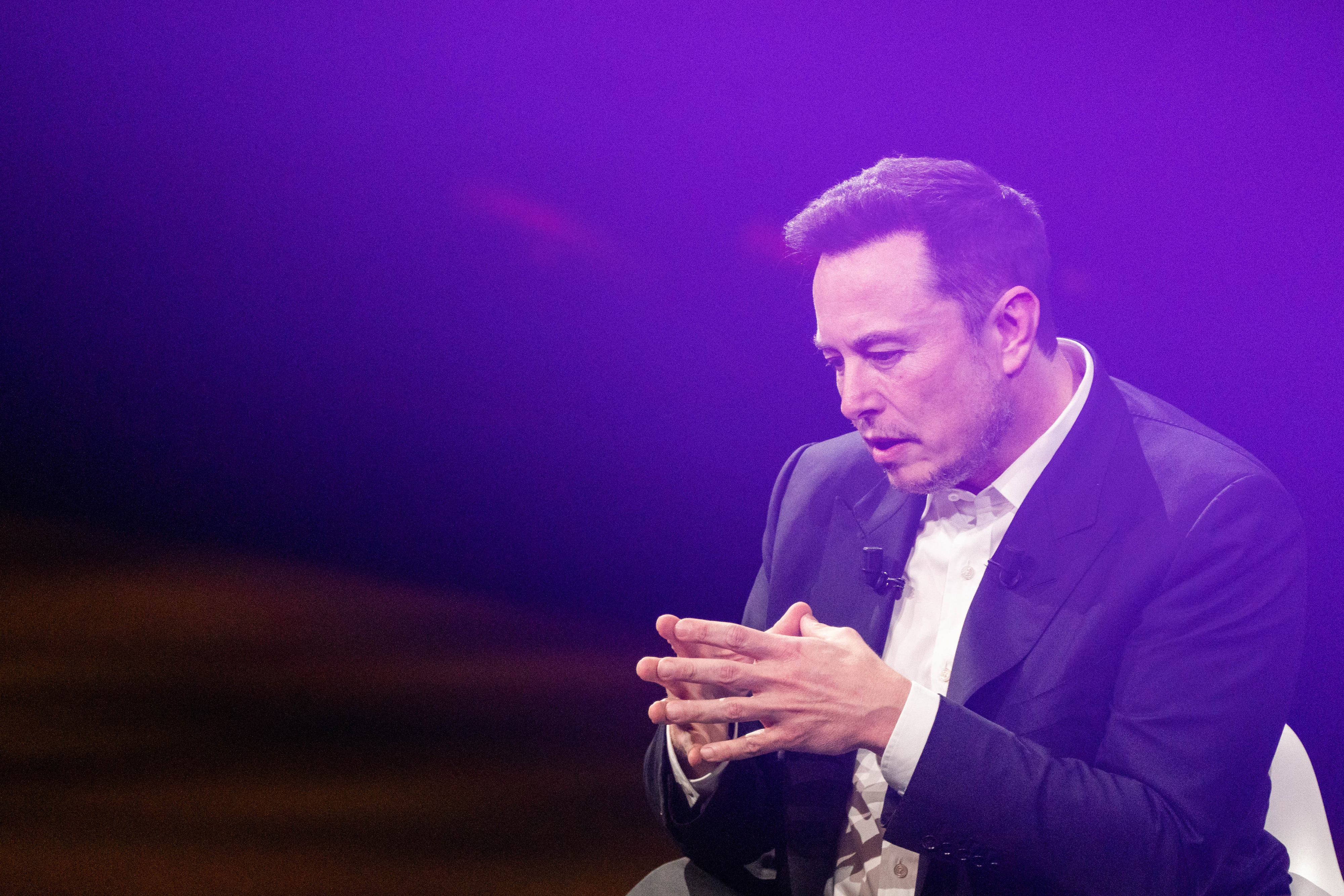 Elon Musk, wearing a black suit and white shirt, speaks as he puts his hands together in front of him, framed by a purple background.