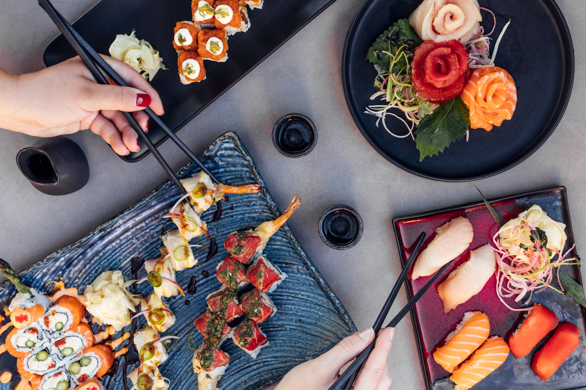 A spread of various sushi dishes, including rolls and nigiri, with two hands and chopsticks.