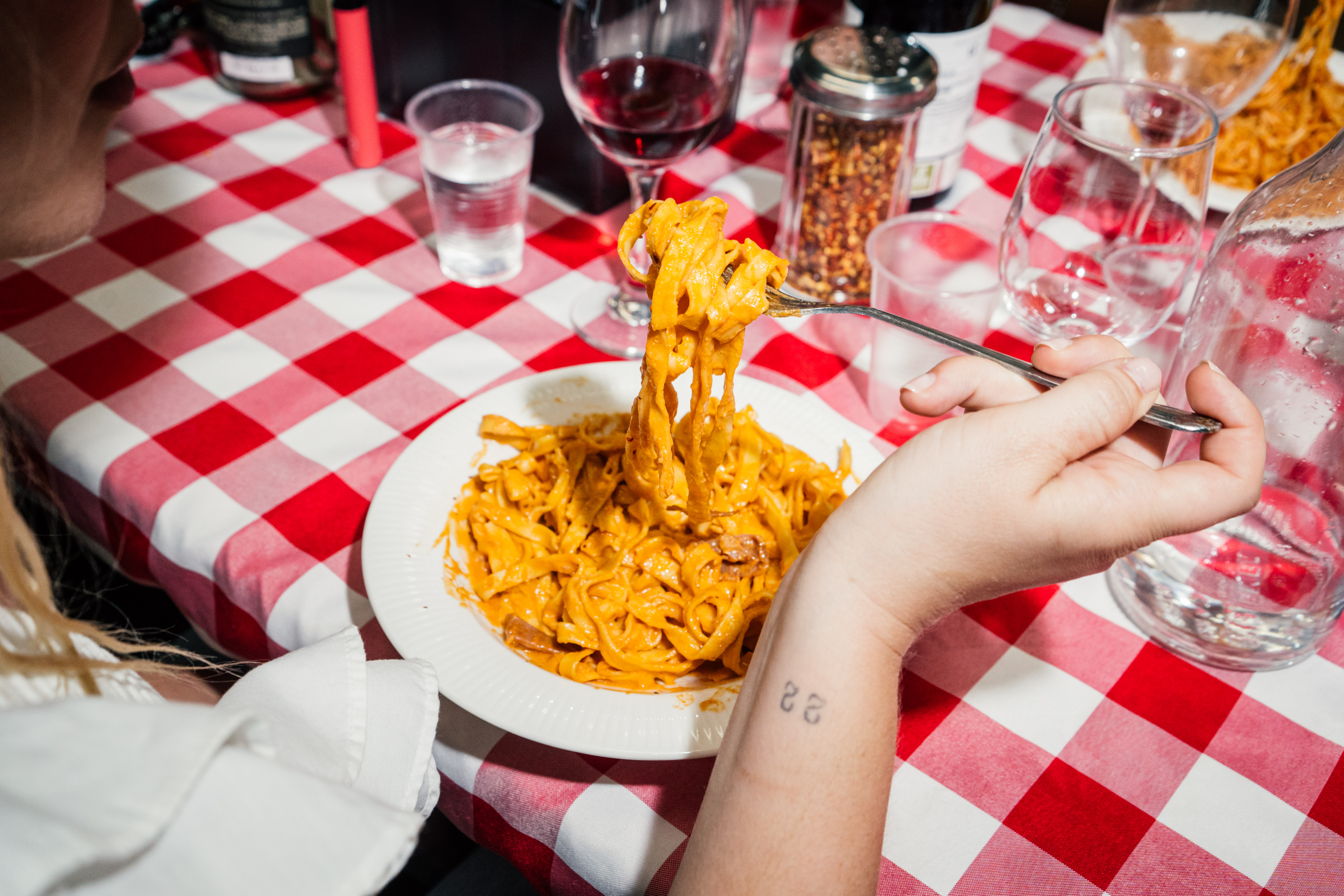 A hand twirls a plate of pasta on a red gingham table.