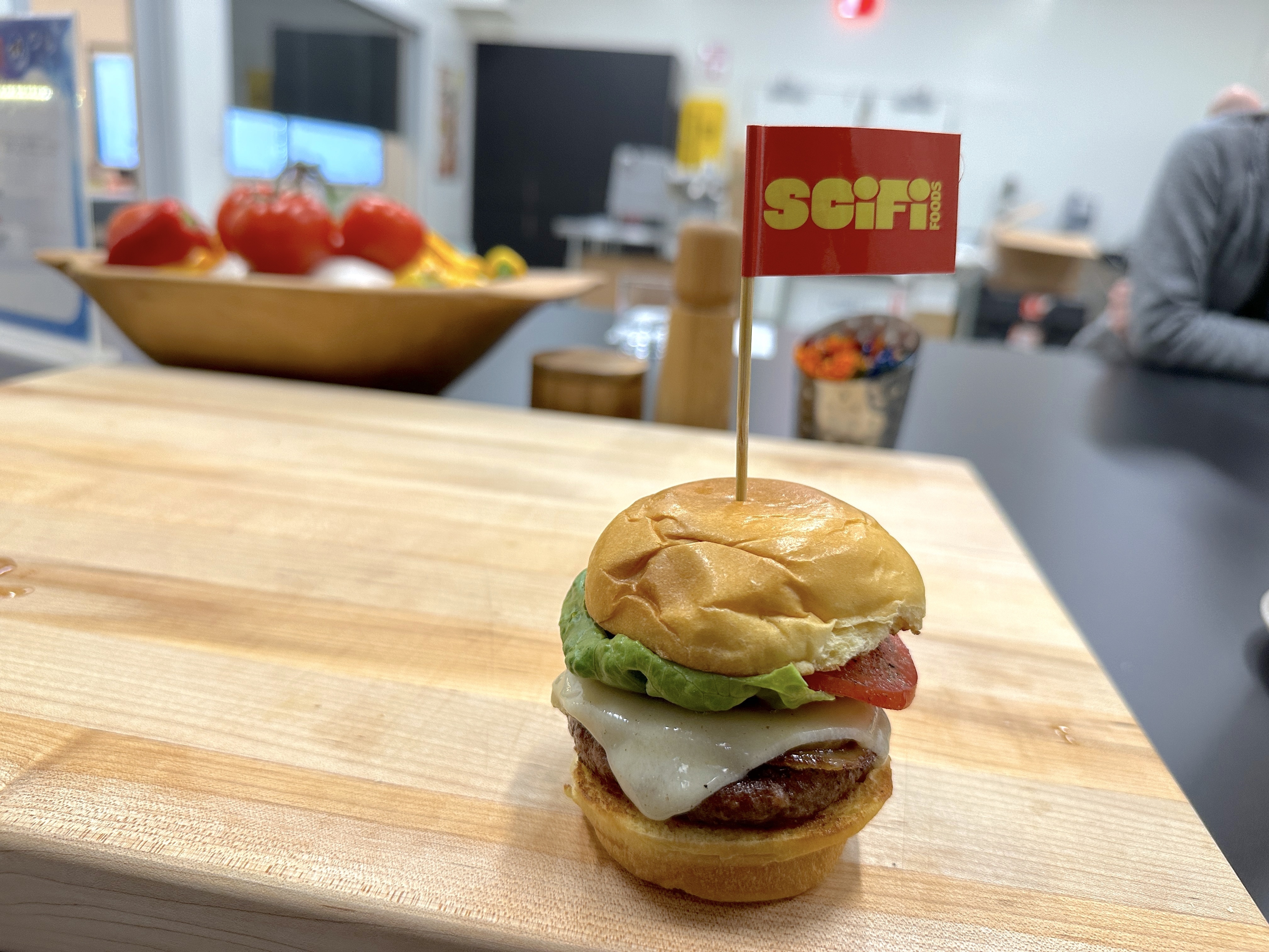 A hamburger with a lab-grown meat patty, along with cheese, lettuce, and tomato, sits on a wooden cutting board. A toothpick with a small red flag affixed to it sticks out of the top burger bun; the flag reads, “SCiFi” in yellow letters.