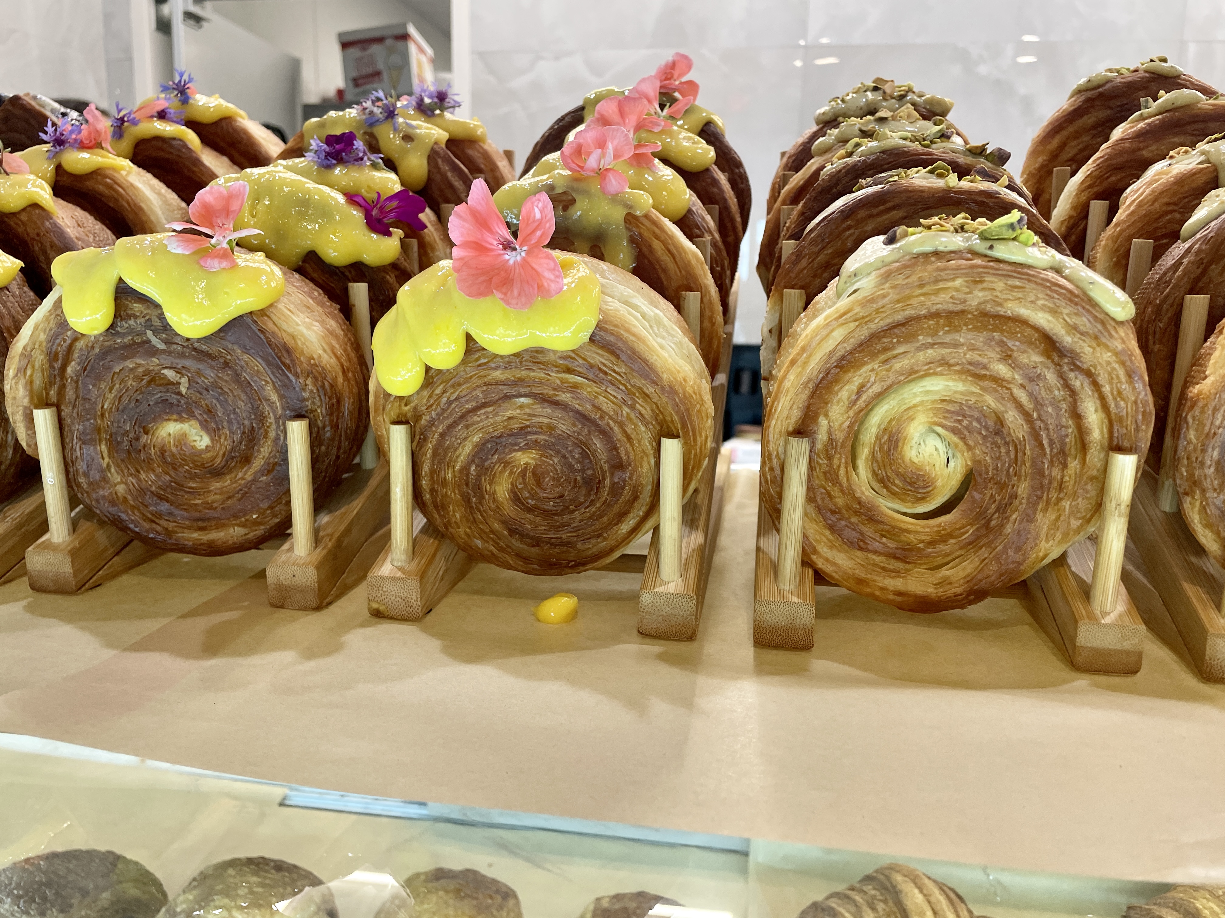 Circle-shaped croissants topped with yellow icing and edible flowers and others with pistachio icing with crushed pistachios sit in wooden holders, facing upright, on a bakery counter.