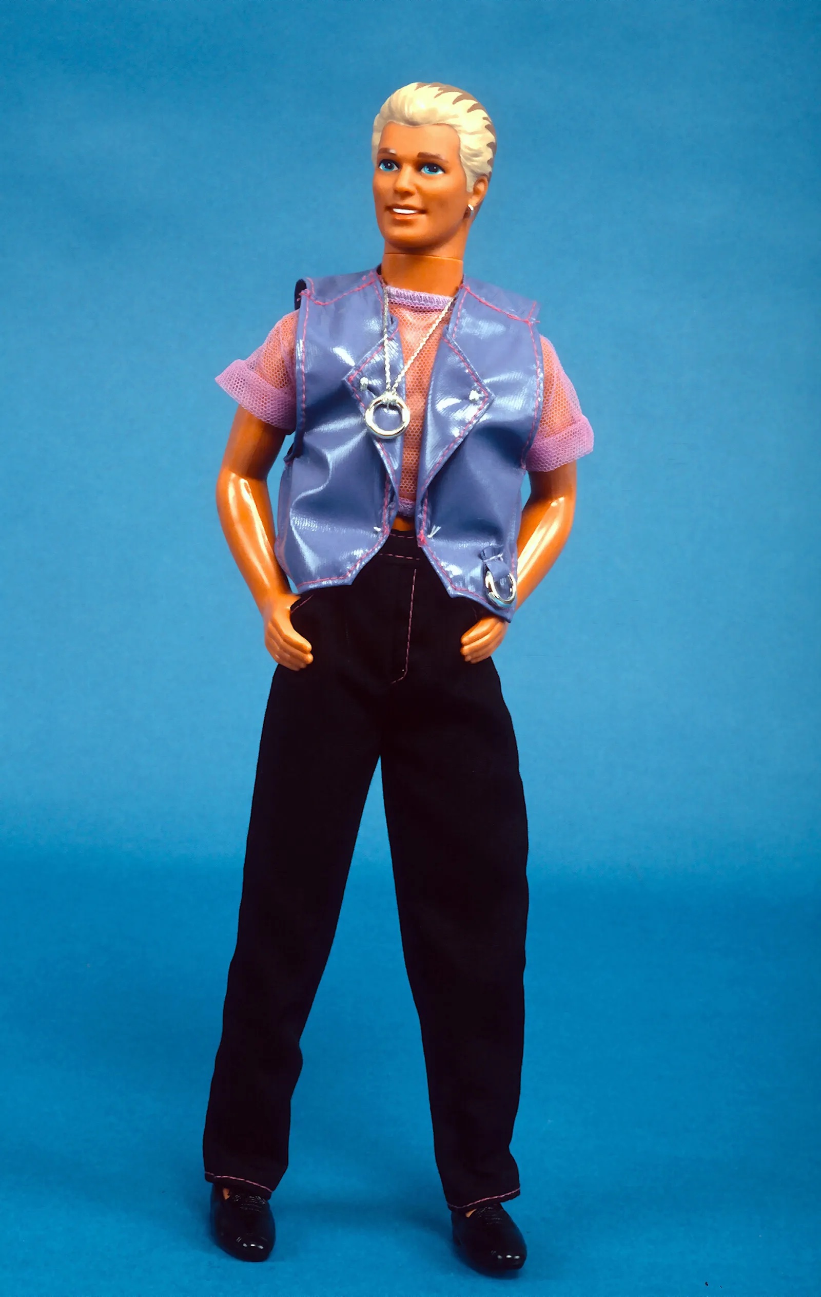 Earring Magic Ken in a glamour shot, hands hooked into his pockets, mesh shirt and shiny pleather purple vest all aglow