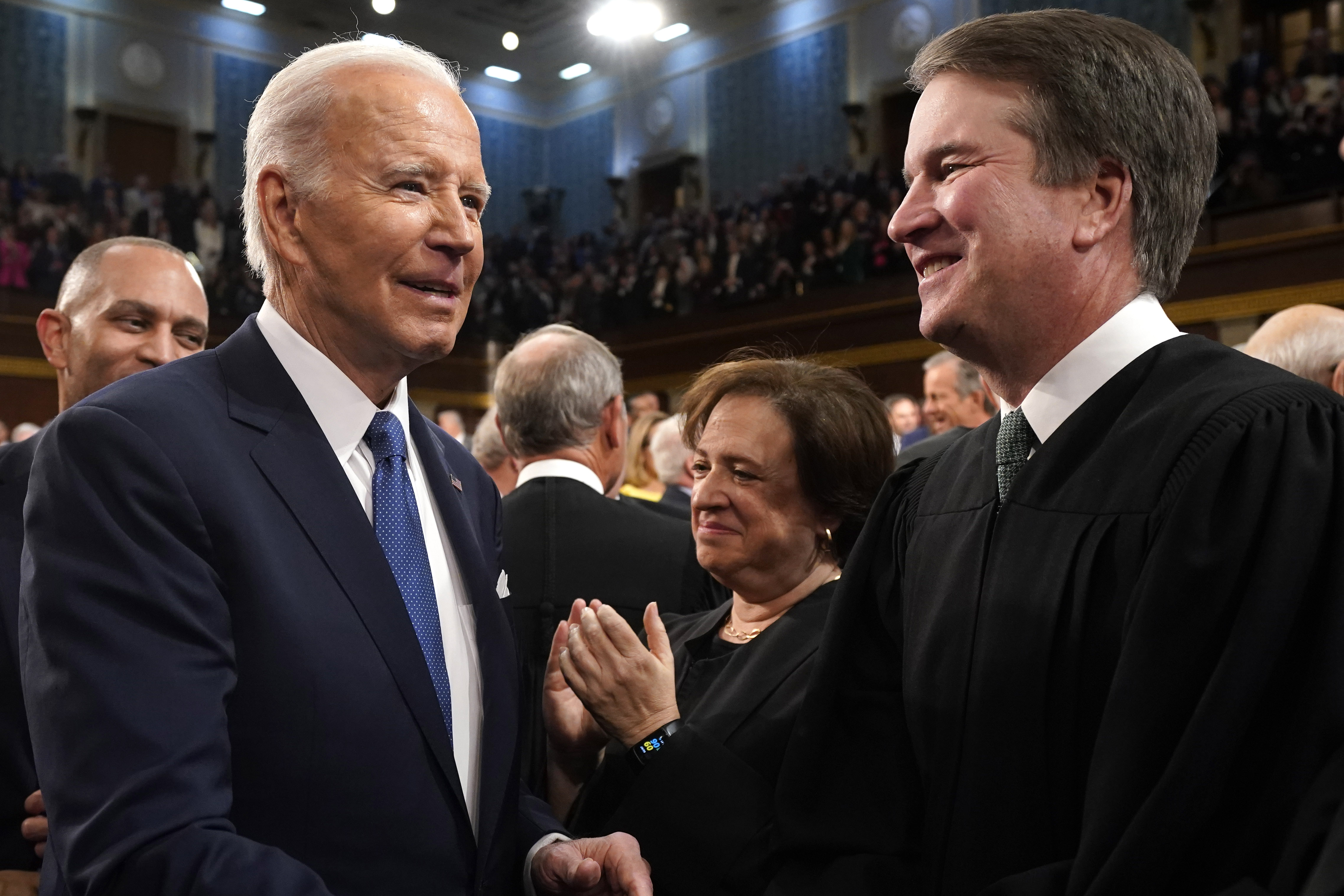 Buden and Kavanaugh face each other, with a crowded room of people in the background. 