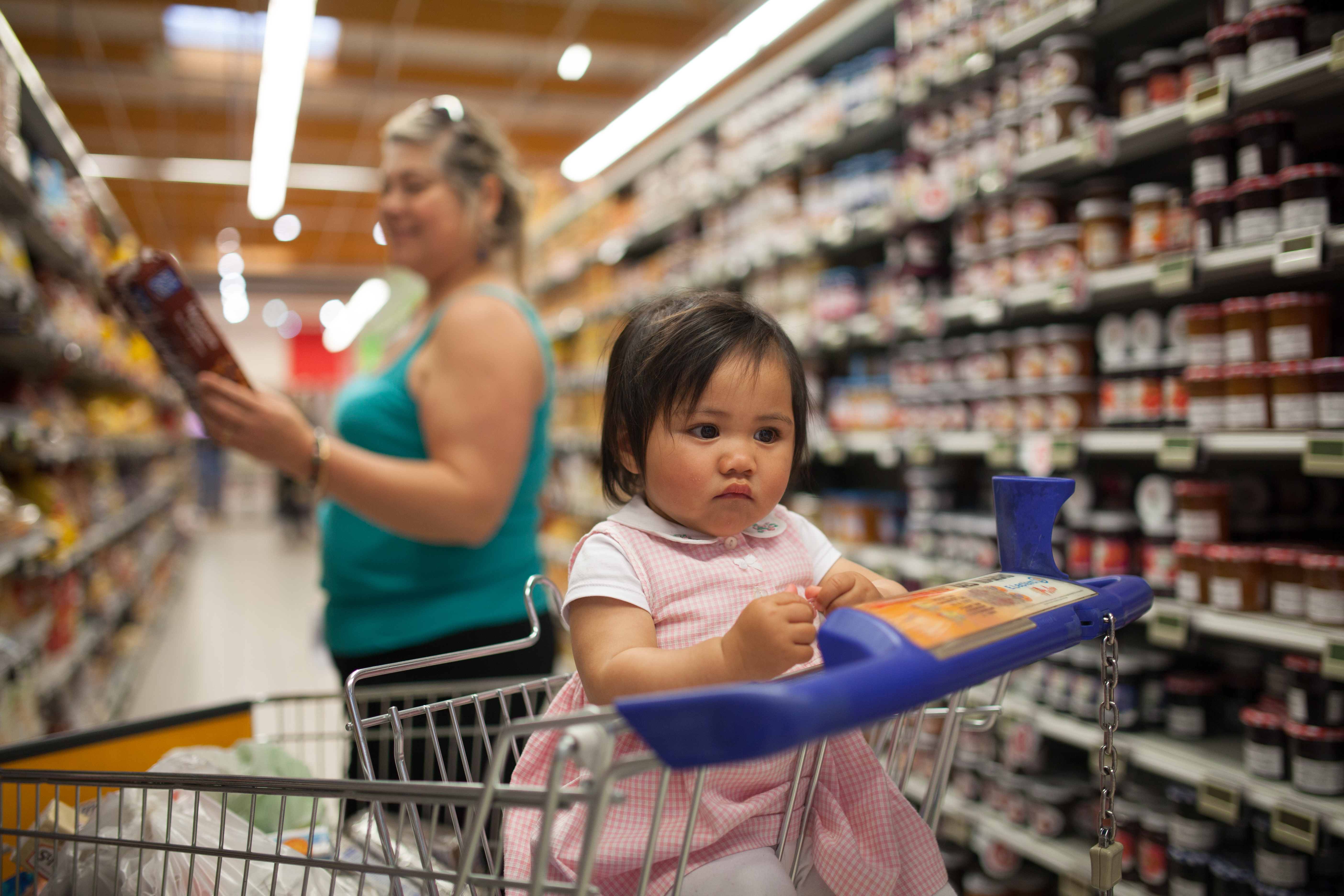 A baby sits in a grocery cart at the supermarket while a woman standing behind the cart examines a product.