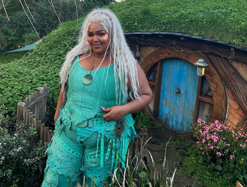 Lizzo wearing a silver wig and green outfit, posing in front of a door in Hobbiton
