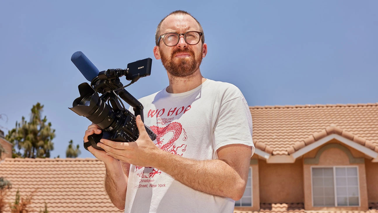 A man with glasses and a beard squints and holds a film camera.