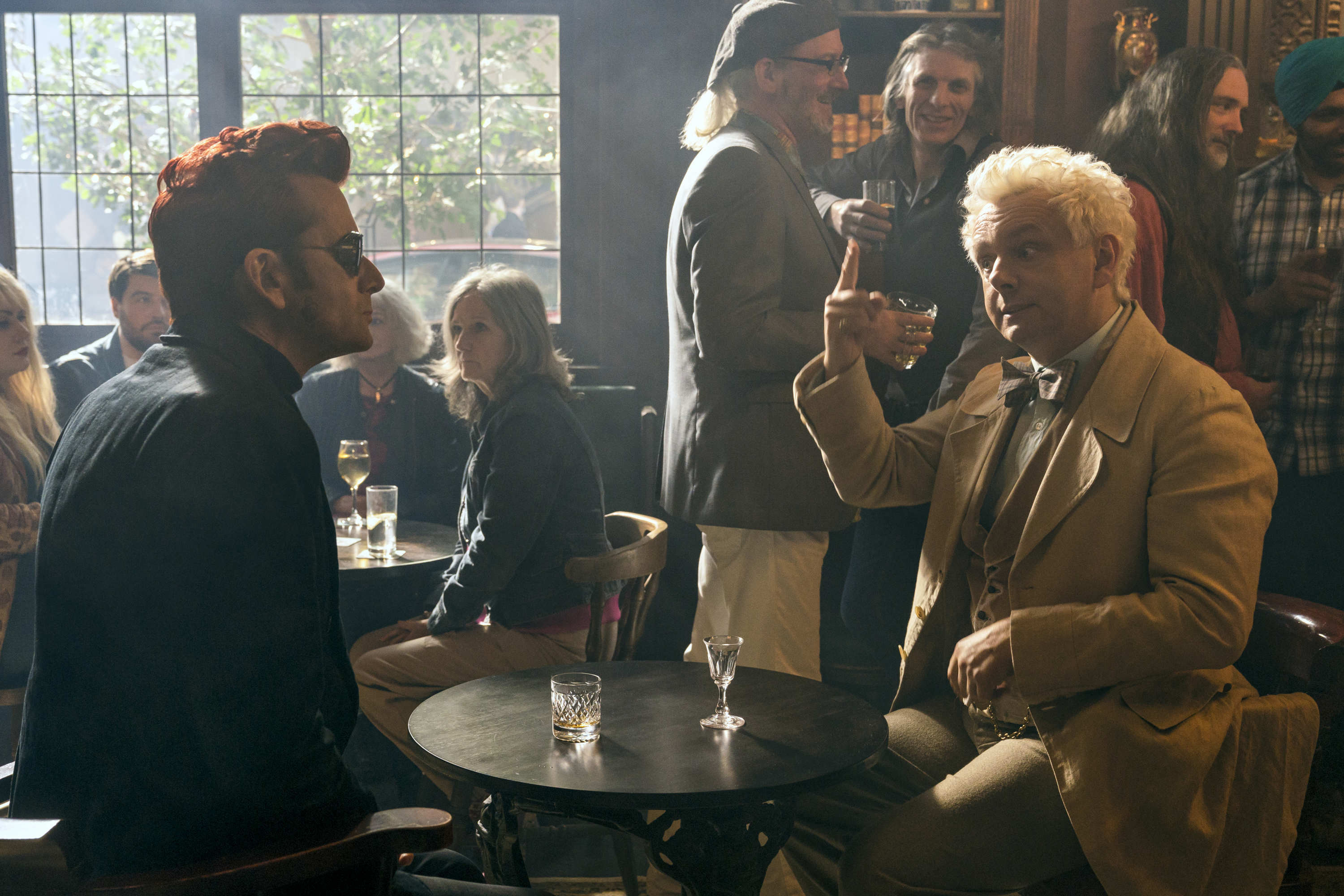 Crowley (David Tennant) sits and looks confused at Aziraphale (Michael Sheen) who is raising his glass to him across the table