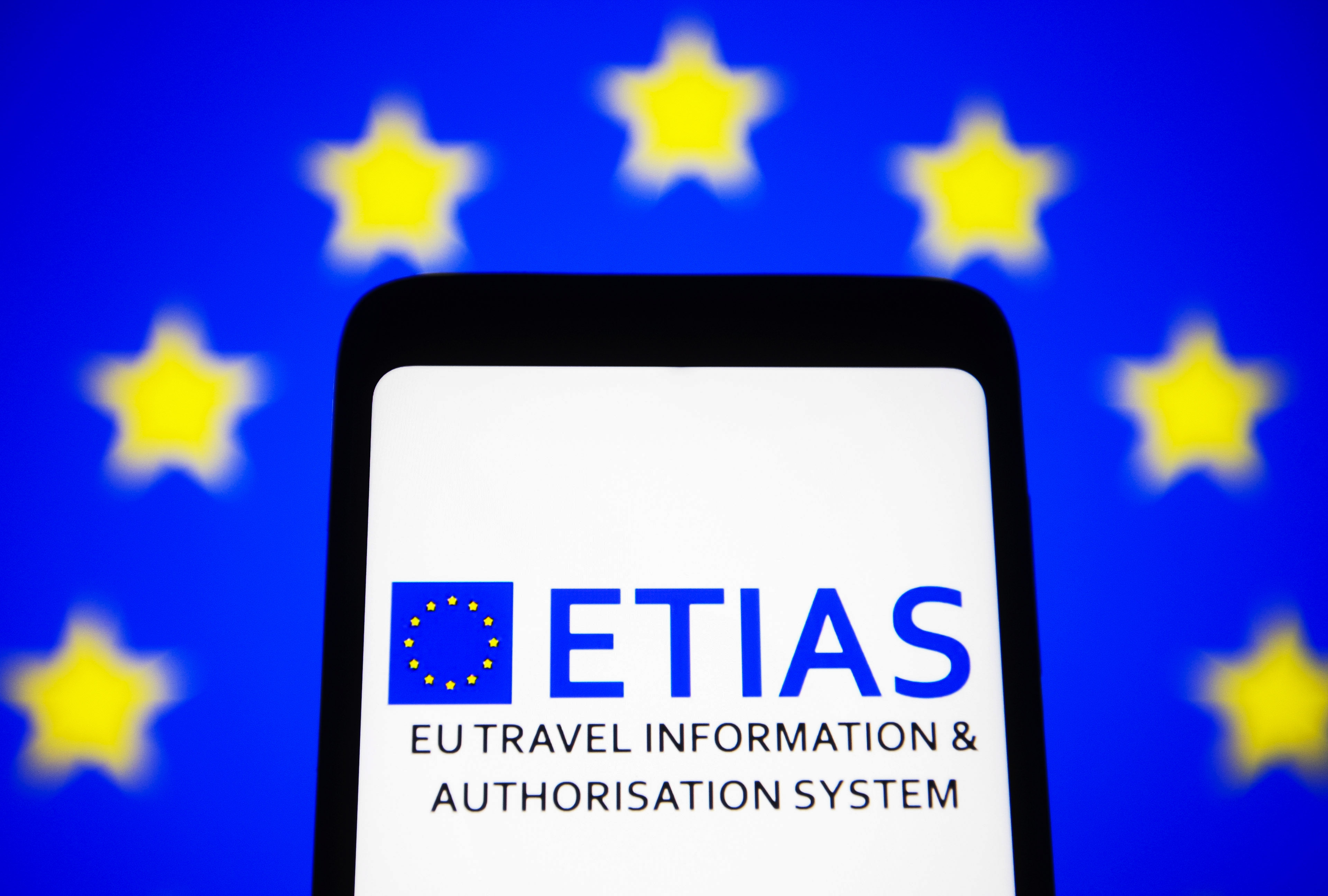 Part of a smartphone screen depicts the ETIAS logo in blue letters, with the European Union flag (blue with yellow stars) in the background.
