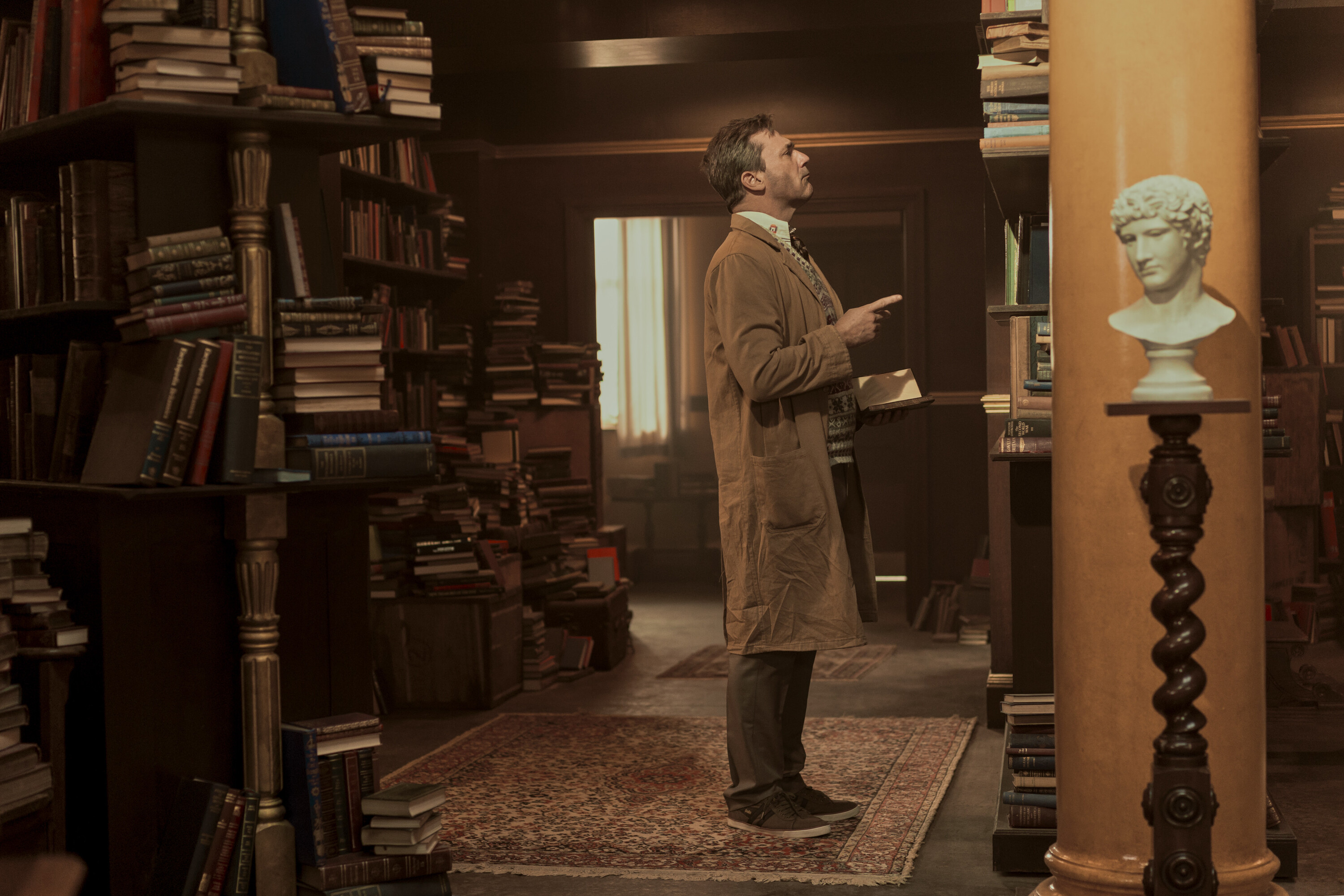 Gabriel (Jon Hamm) standing in front of a bookshelf and pointing like he’s looking for where to put a book
