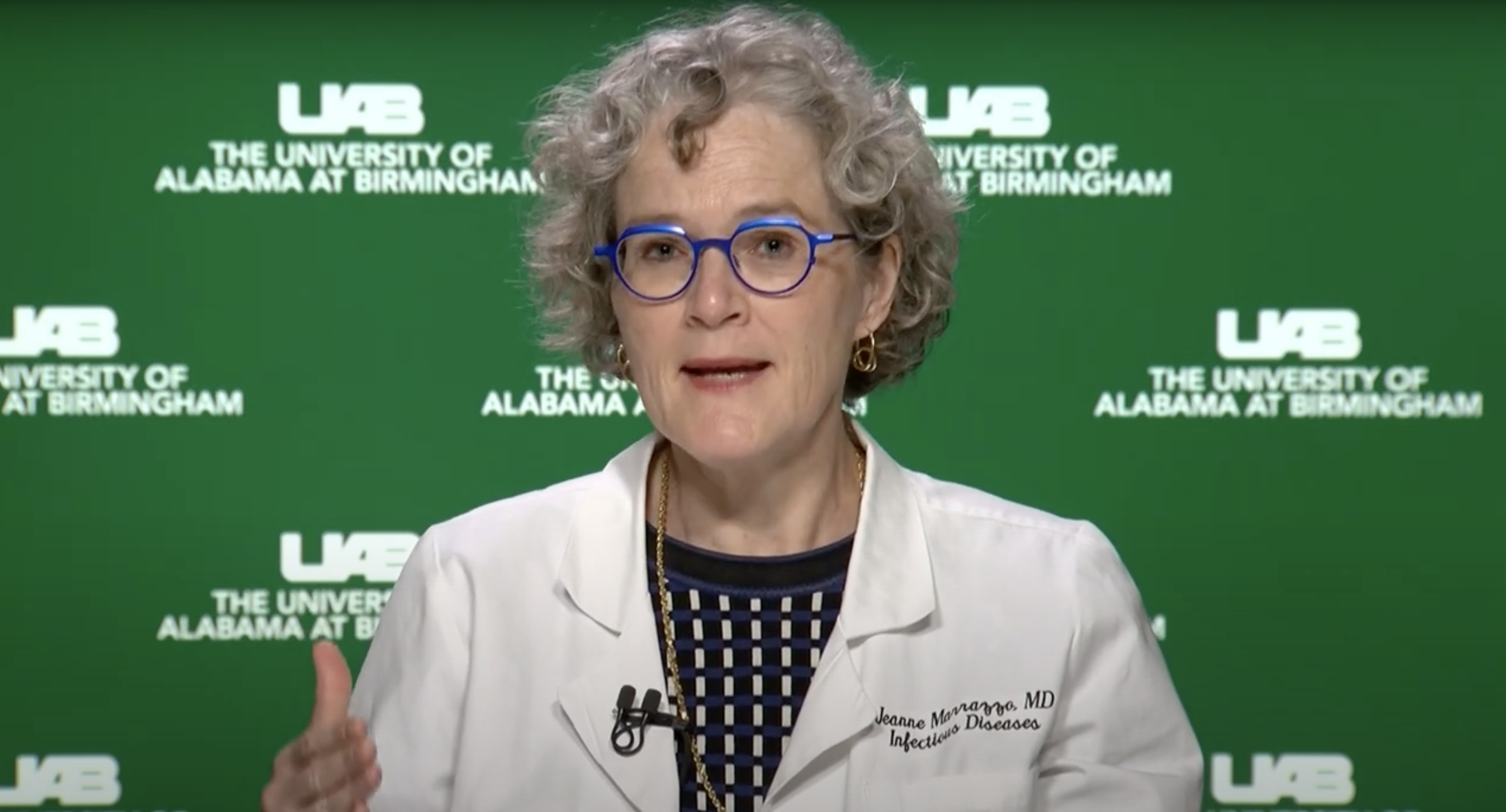 A white woman with glasses speaks against a green and white backdrop.