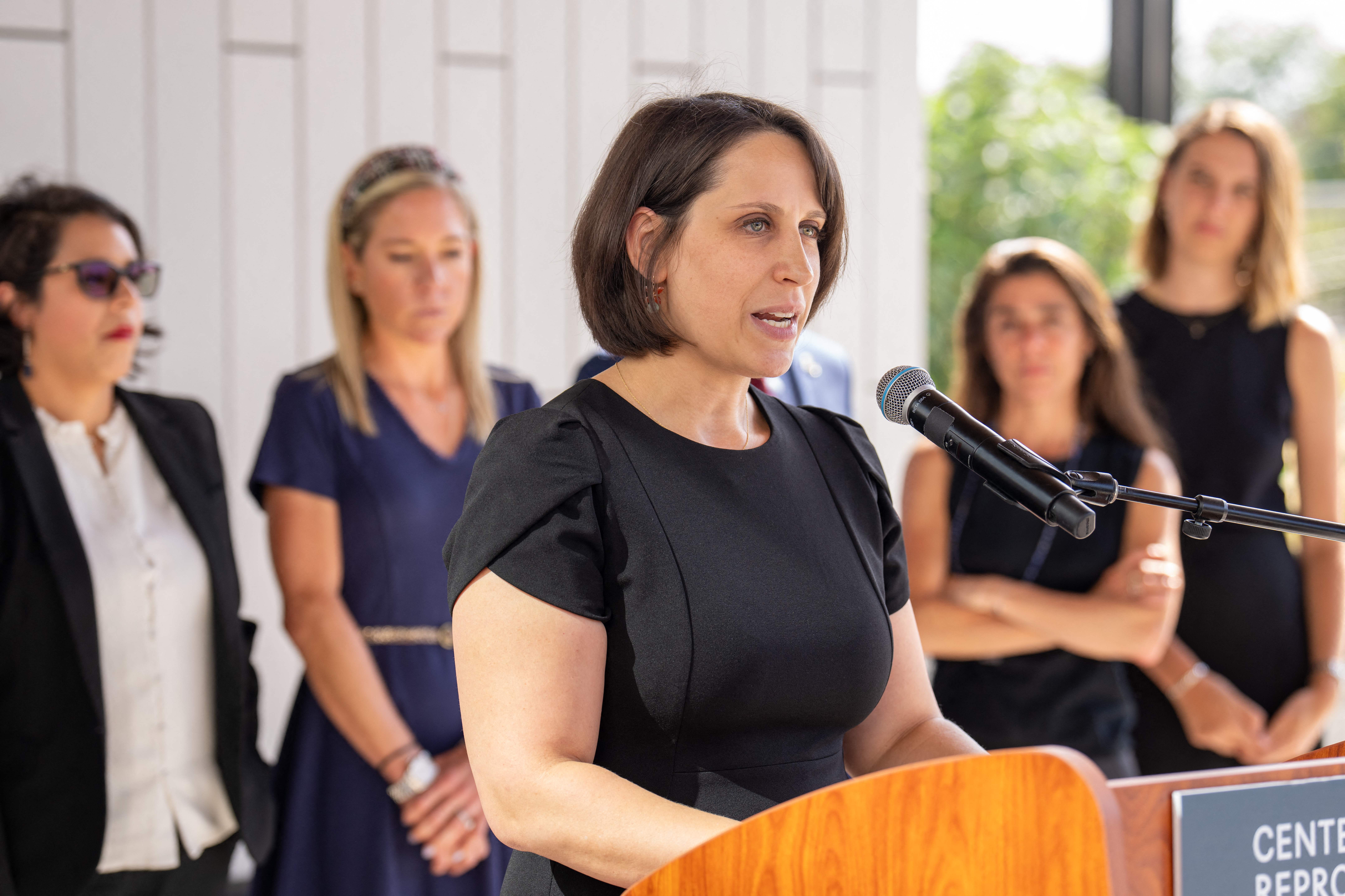 Molly Duane stands before a wooden podium and speaks into a microphone, wearing a black top, and flanked by several other women. 