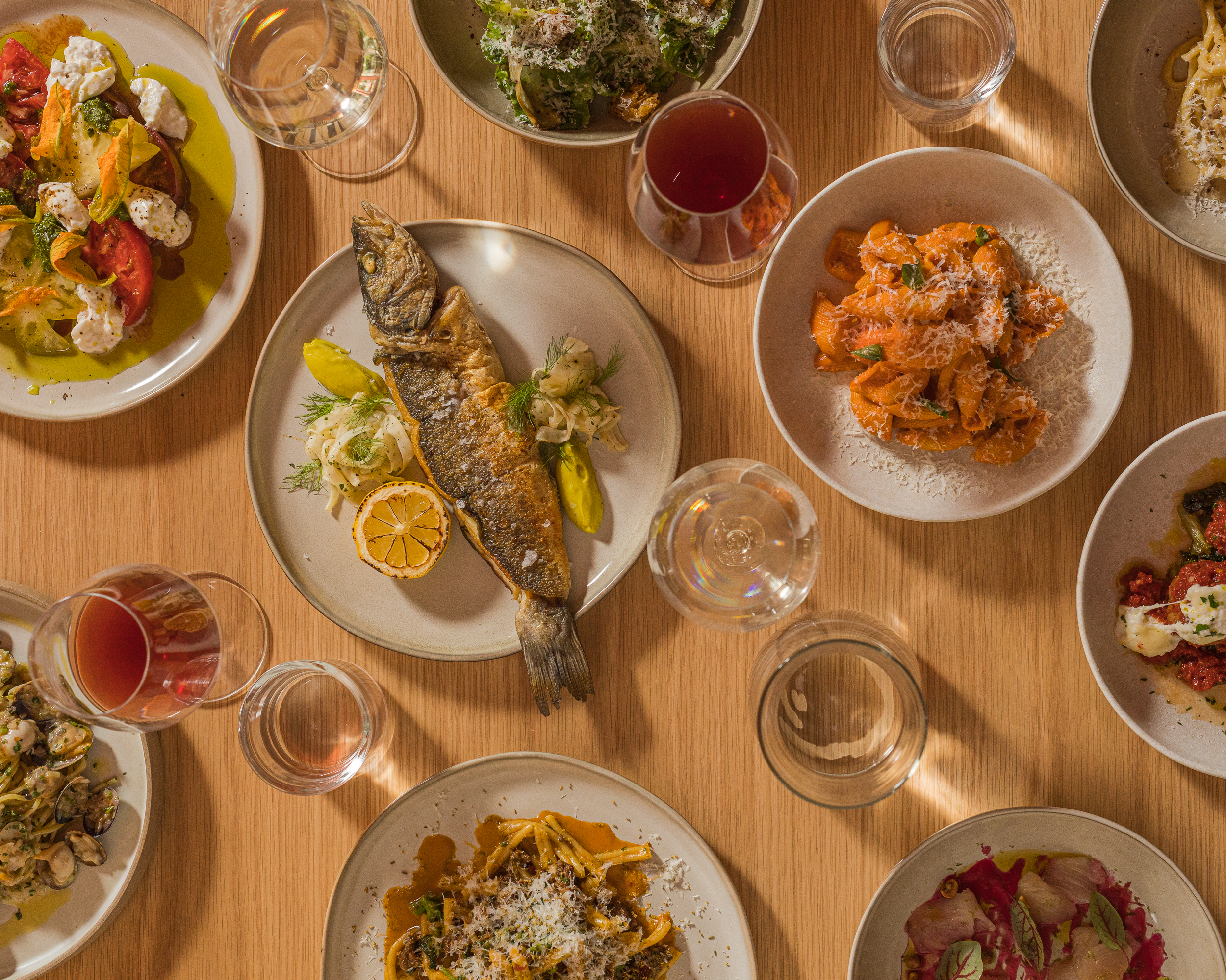 An overhead shot of a light wooden table with whole fried fish, pastas, wine glasses, and more at Mona Pasta Bar.