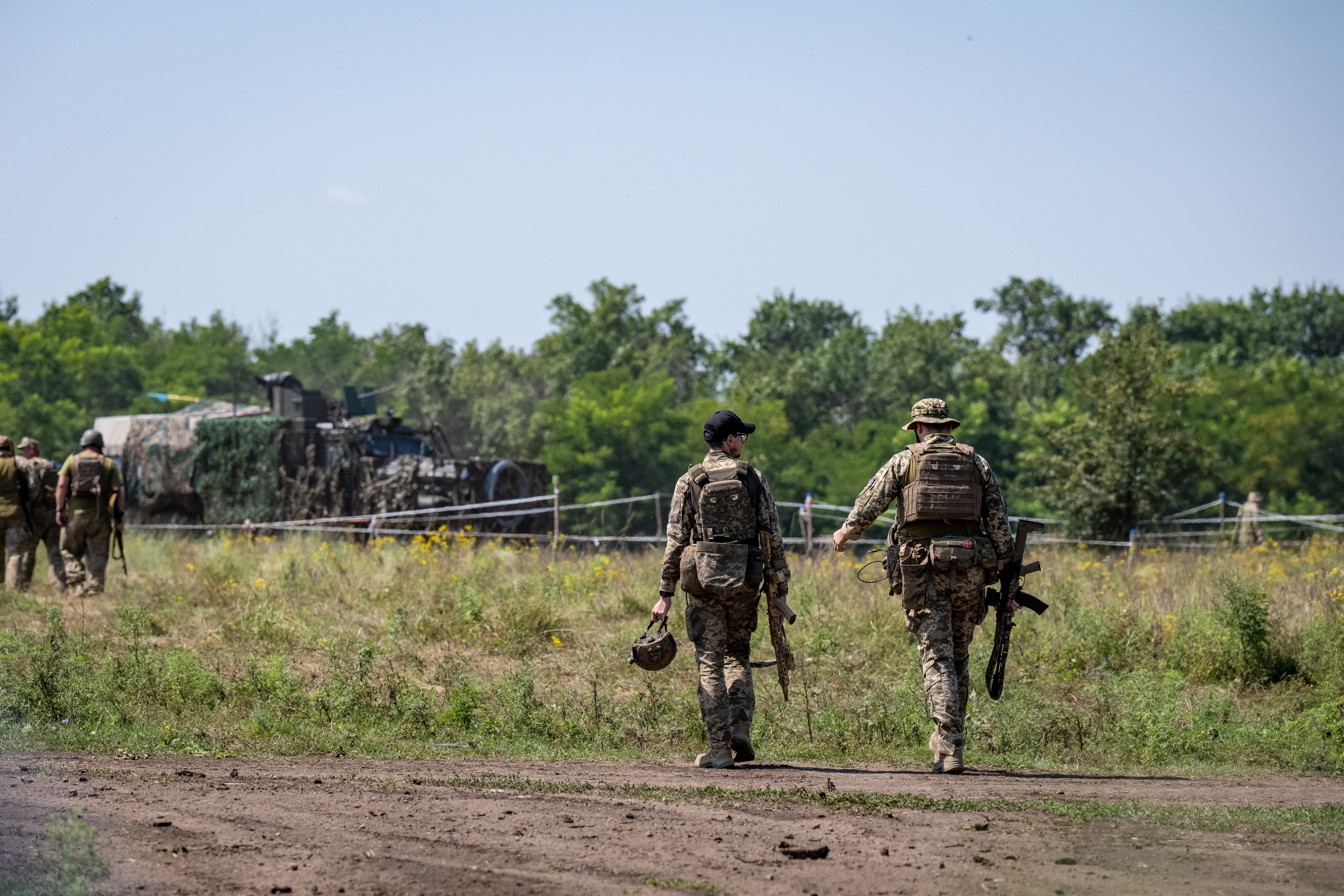 Soldiers walk in a field. A tank is seen in the distance. 