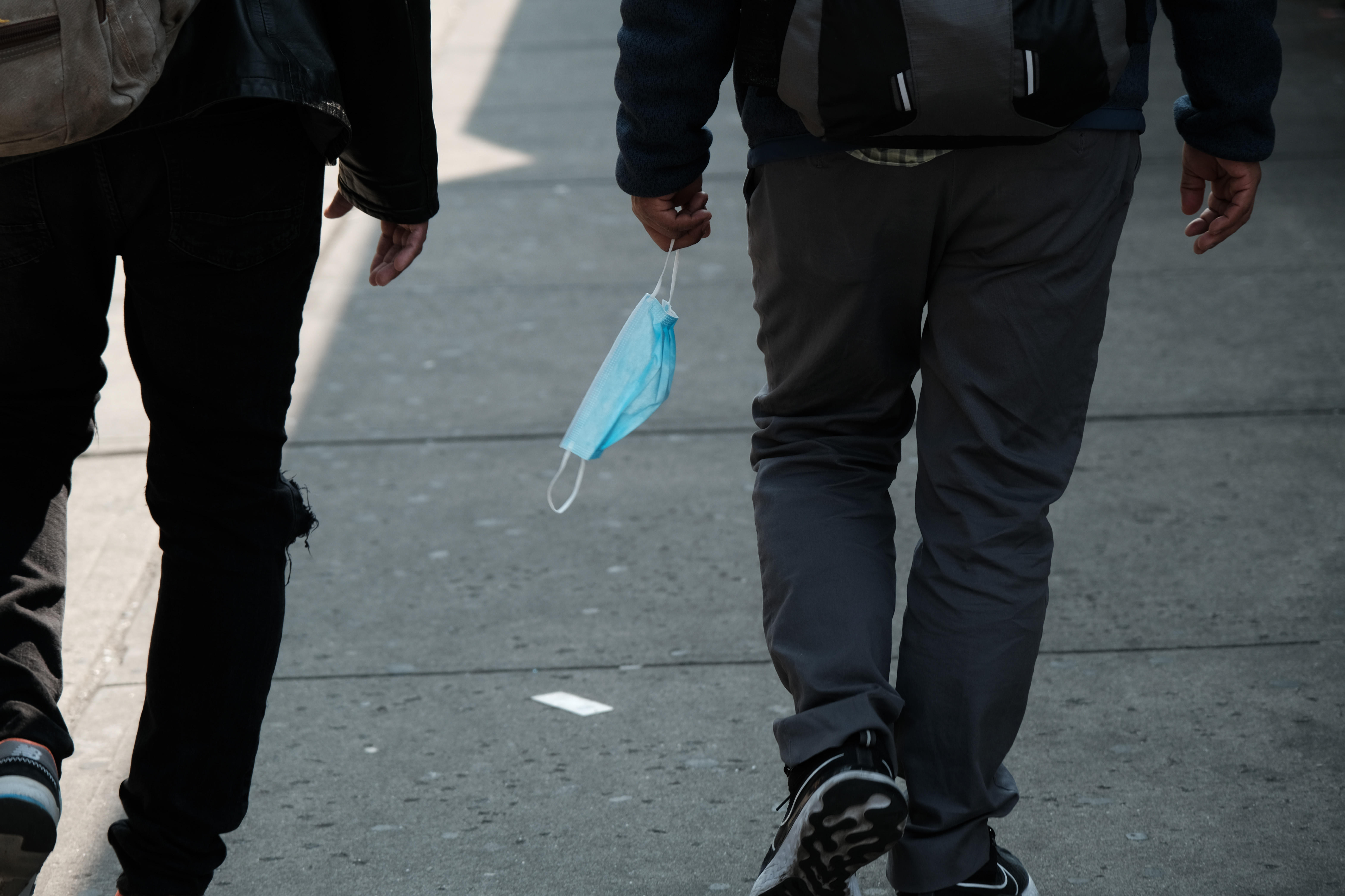 People walking on a sidewalk, one dangling a disposable face mask from their hand.