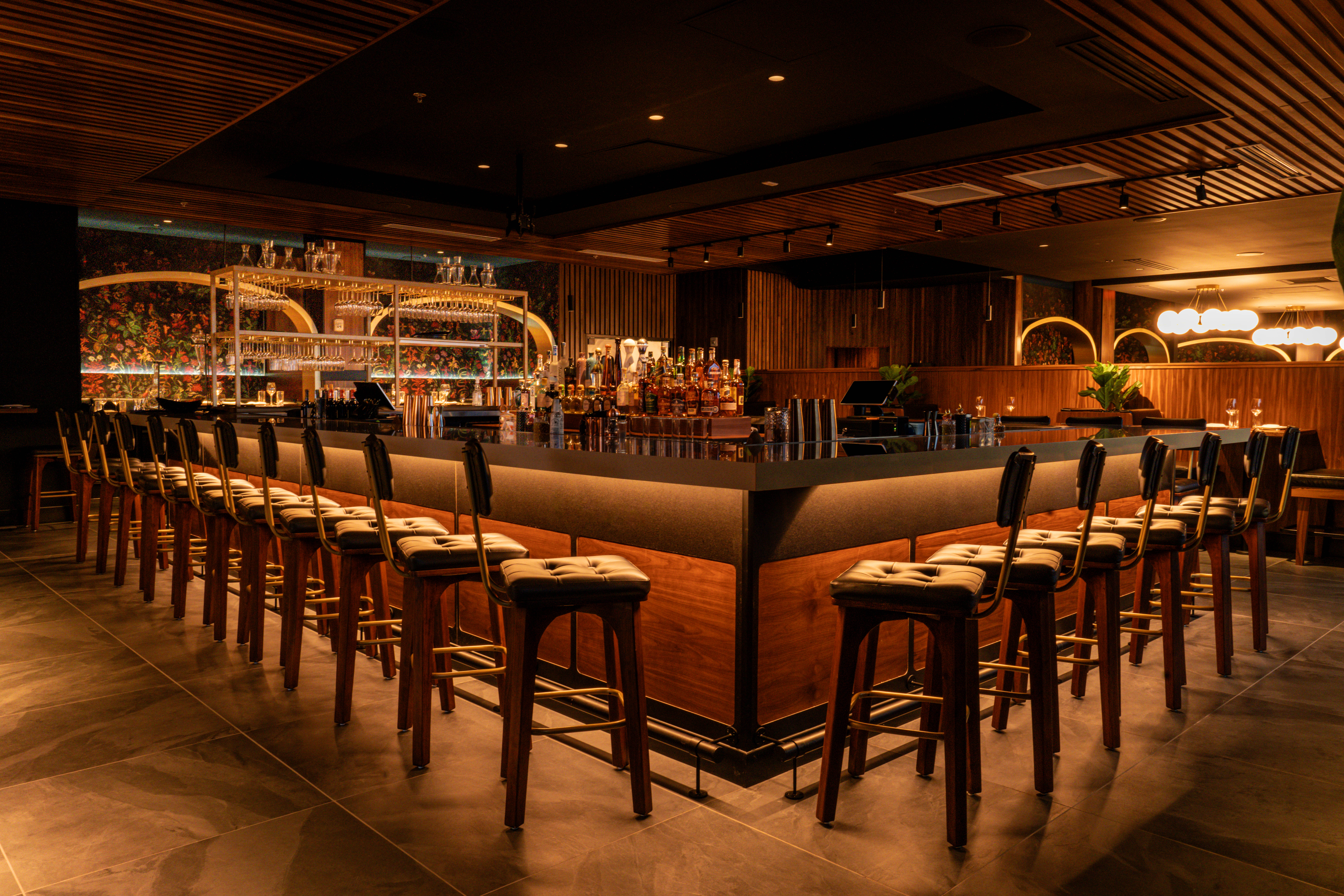 A rectangular wood-paneled bar surrounded b tufted stools a full wall of liquor bottles towards the back. 