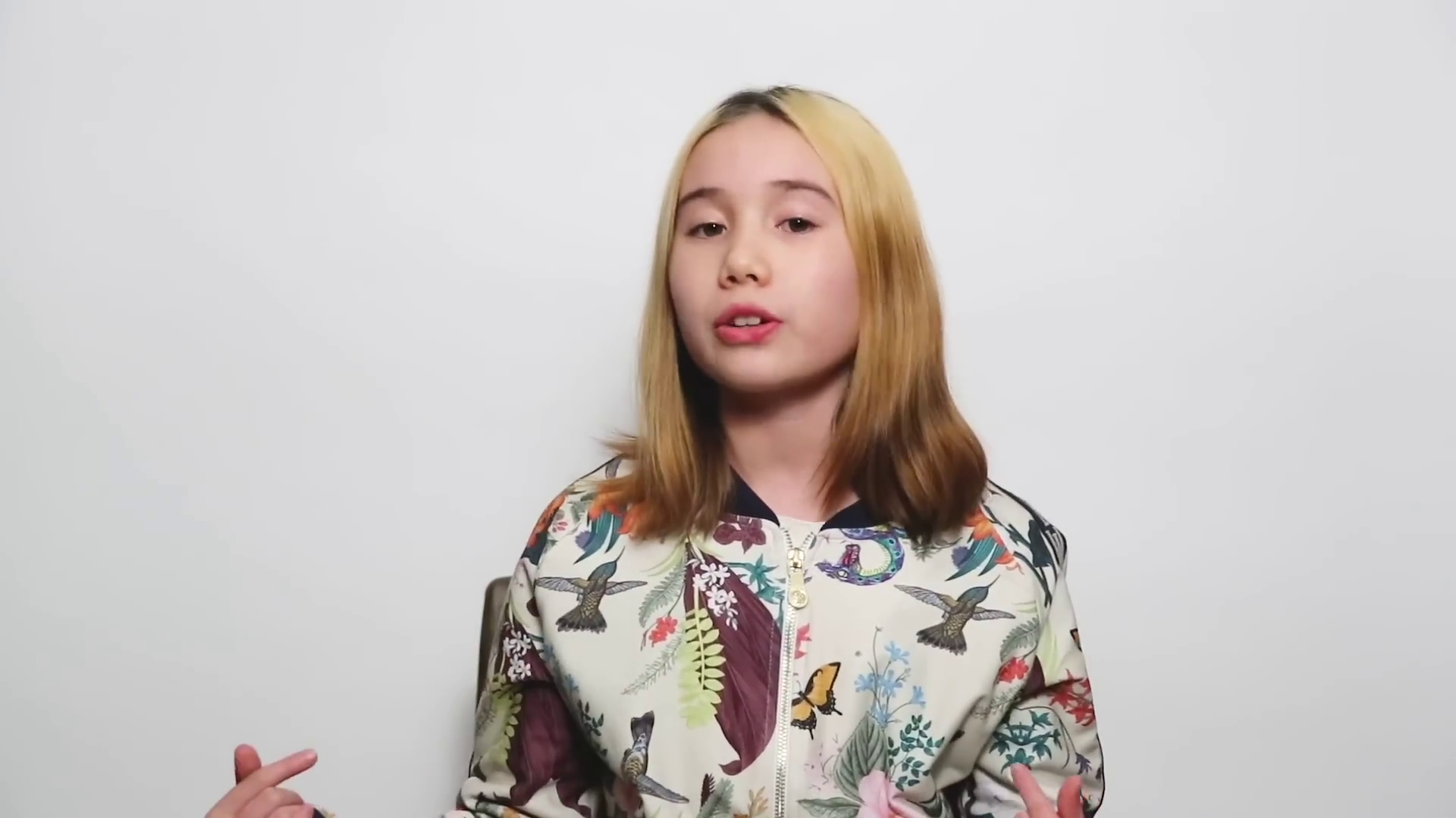 A still from a video of Lil Tay. She’s sitting in front of a white background. She’s younger in this video, around age 9 or 10. She’s looking off into the camera and gesturing with her hand. 