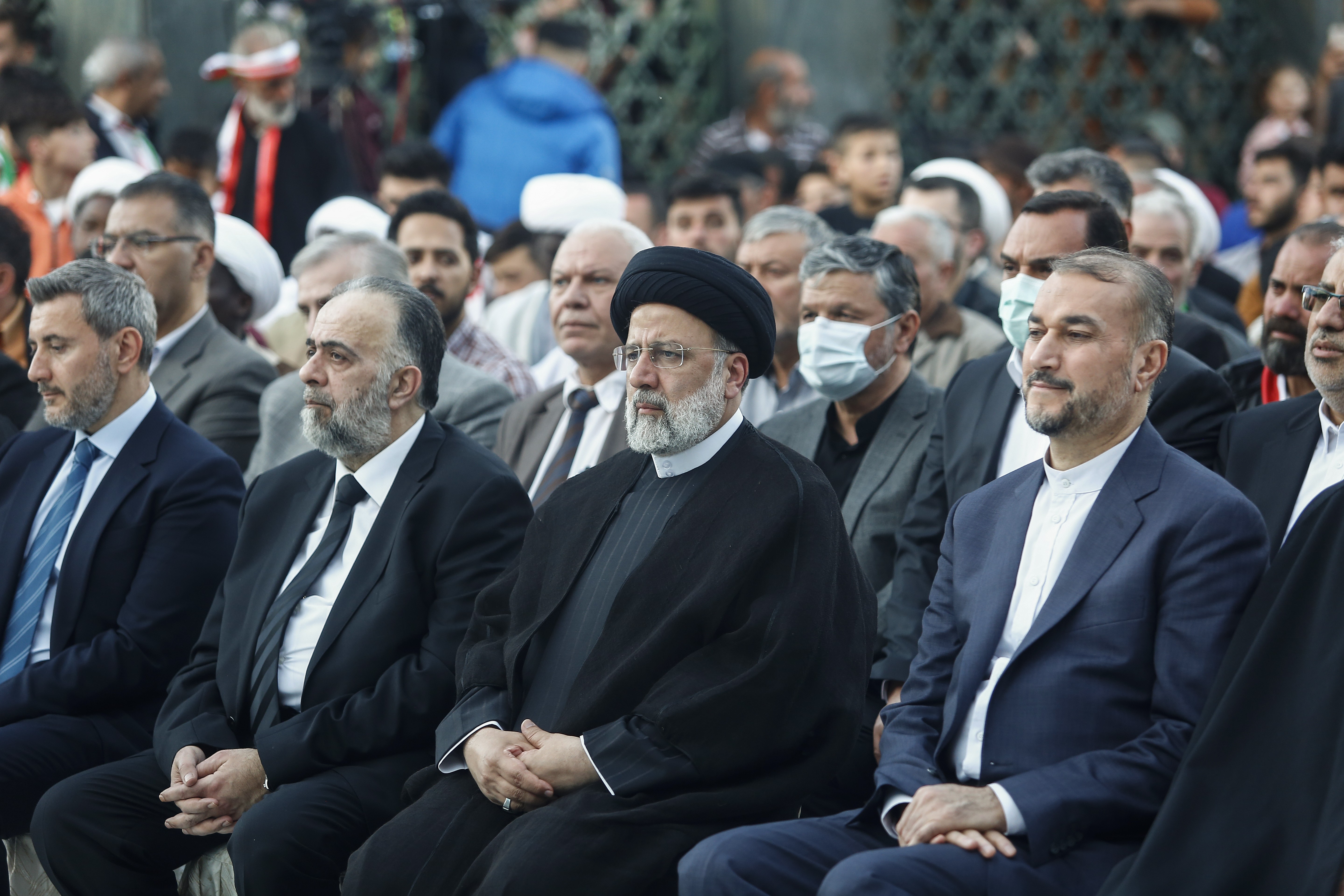 Men wearing suits with hands clasped in their laps are seated in rows. President Ebrahim Raisi sits in the front row and wears a black turban on his head. 