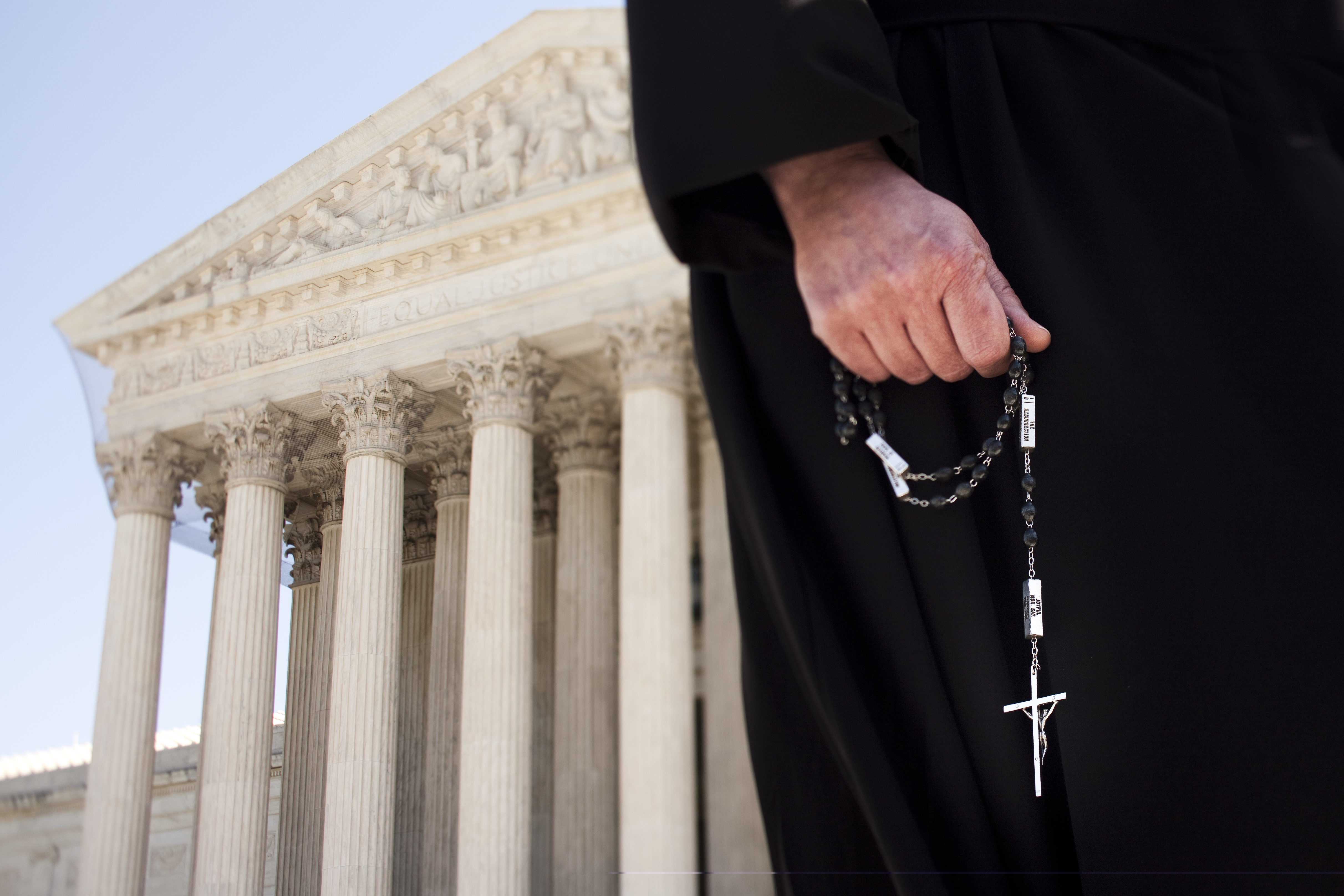 A person in a black robe holding a religious cross on a chain. The Supreme Court building is in the background.