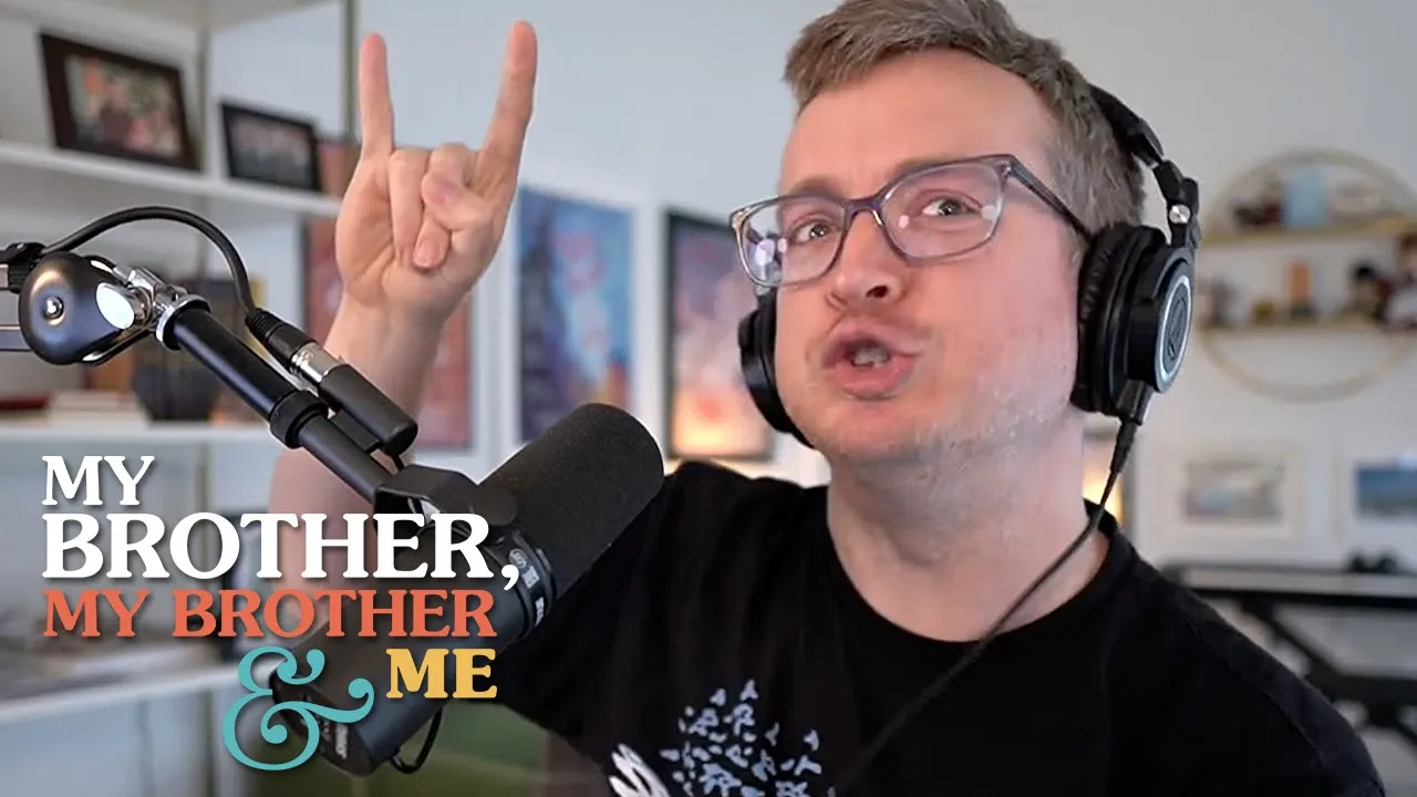 A photo of Griffin McElroy, throwing up the ‘rock on’ horns on his right hand. He is seated in his office, behind a microphone, and he is wearing headphones and glasses. The MBMBaM logo is superimposed on the lower left corner of the image.