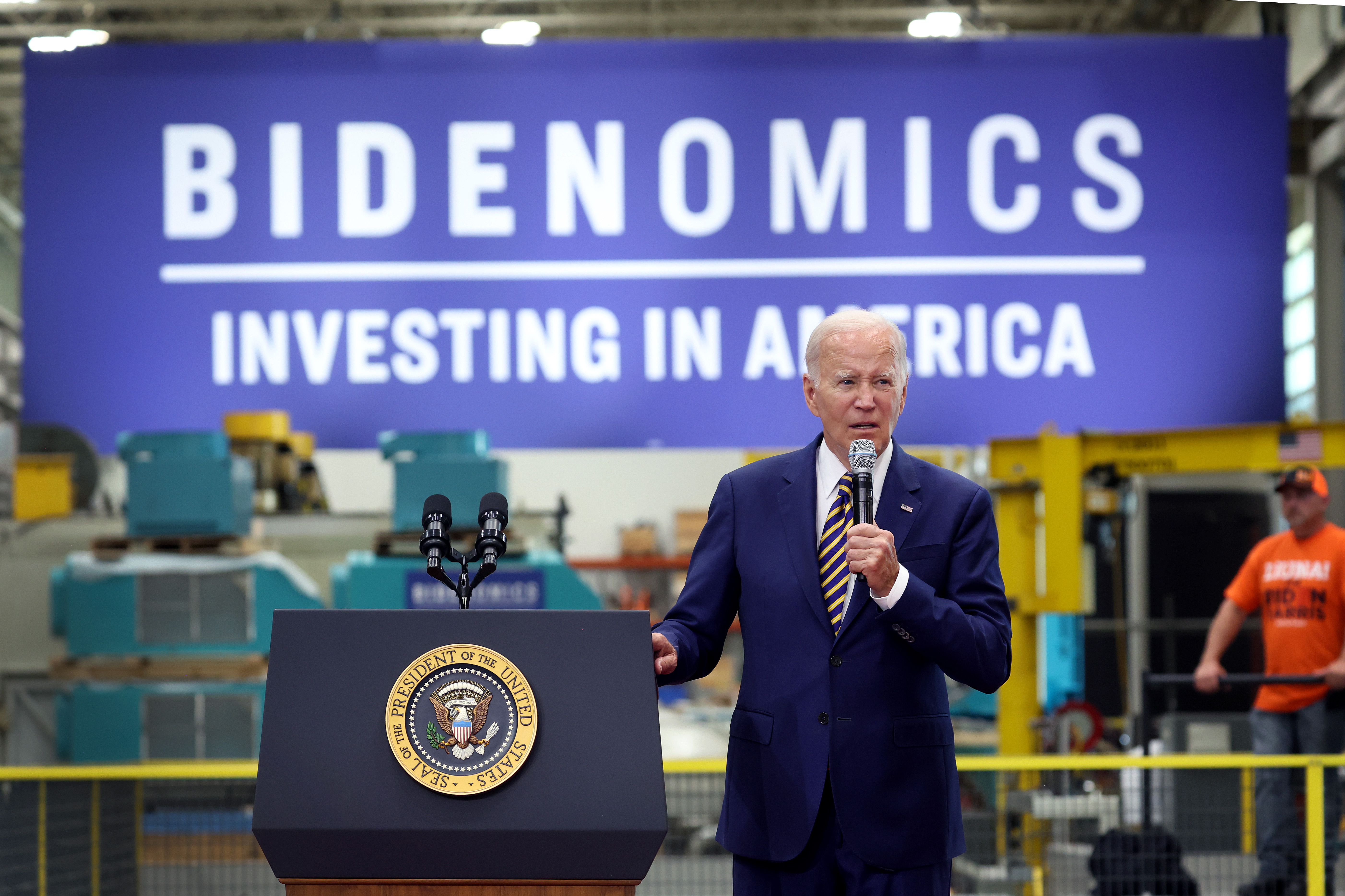 President Biden at an electrical equipment plant speaking beside a lectern in front of a sign that reads “Bidenomics: Investing in America.”