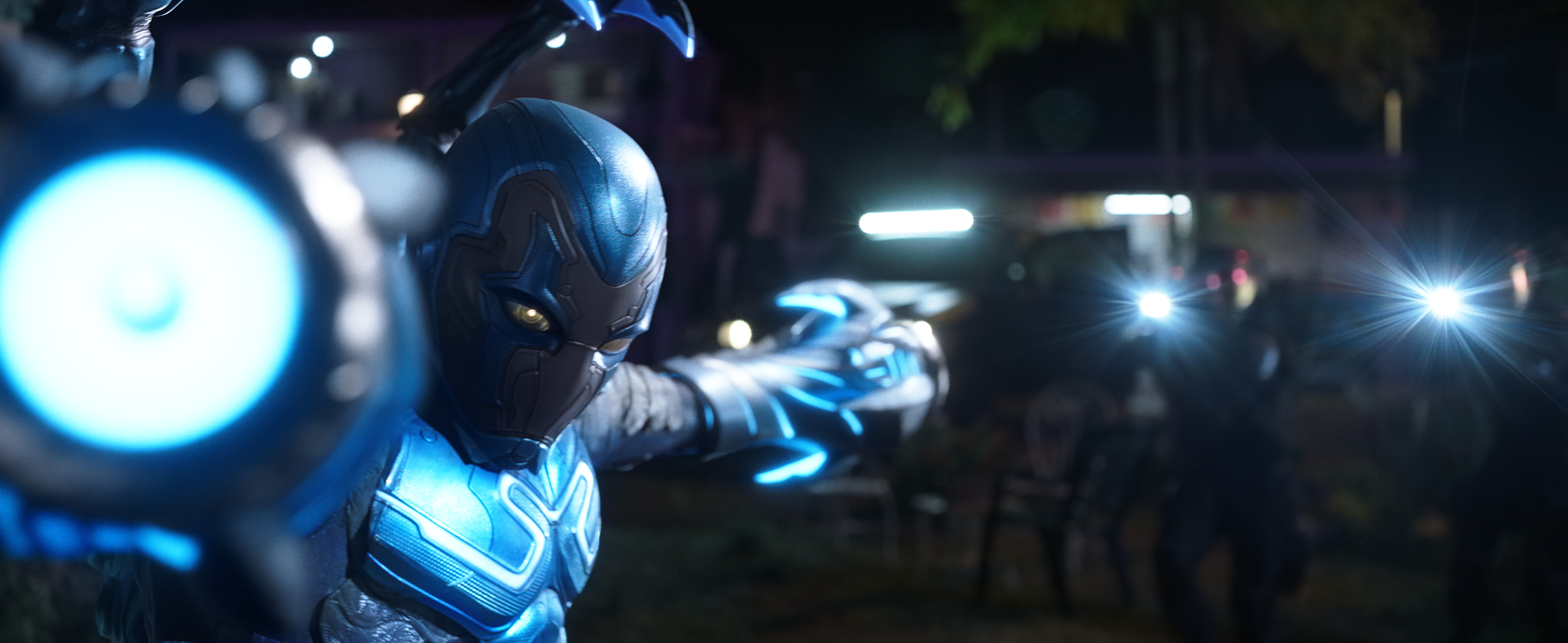 Blue Beetle poses with his arms in the shape of laser canons pointing both directly at and away from the camera in the film Blue Beetle.