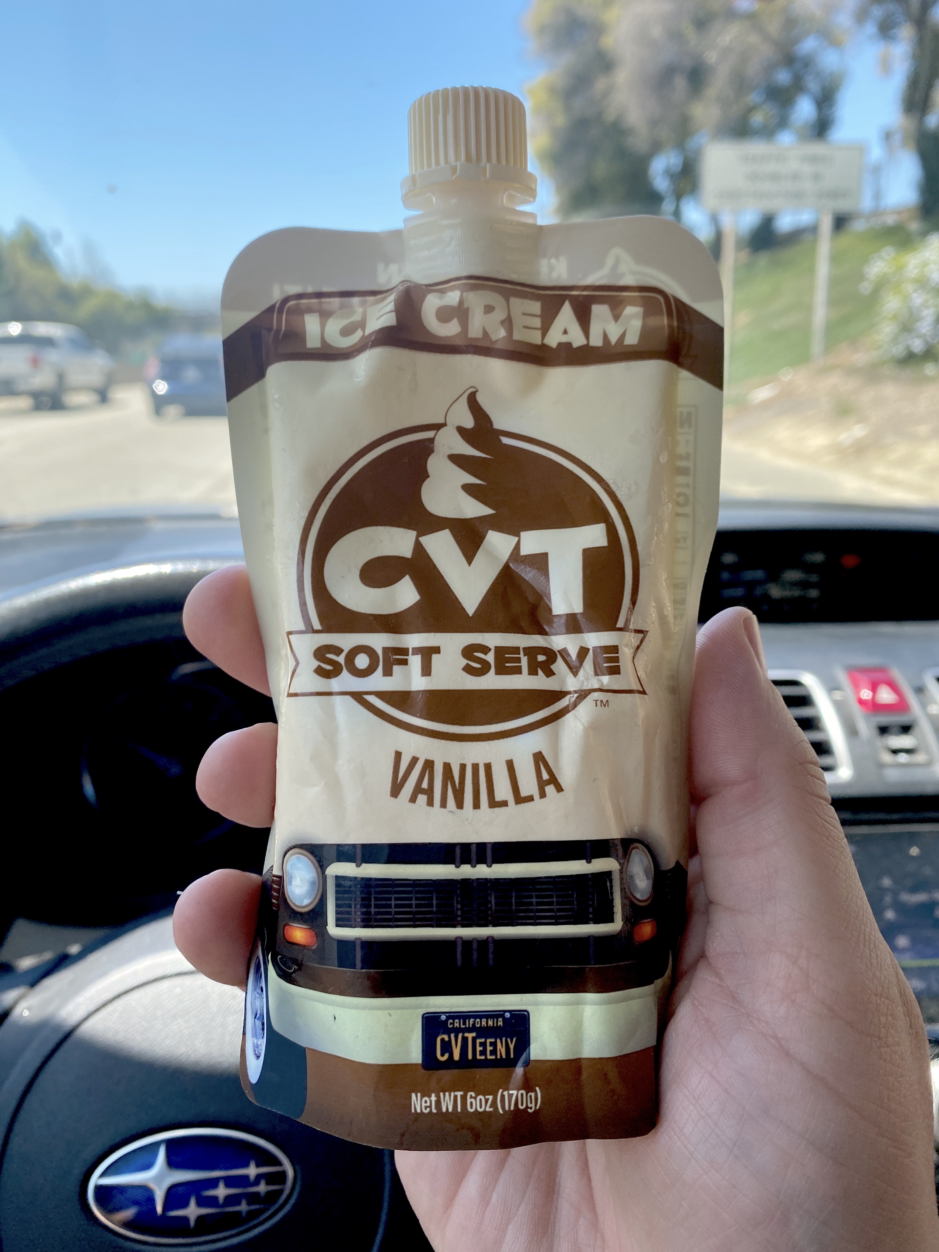 A hand holds a squeeze bag of brown and white soft serve while in a car at daytime.
