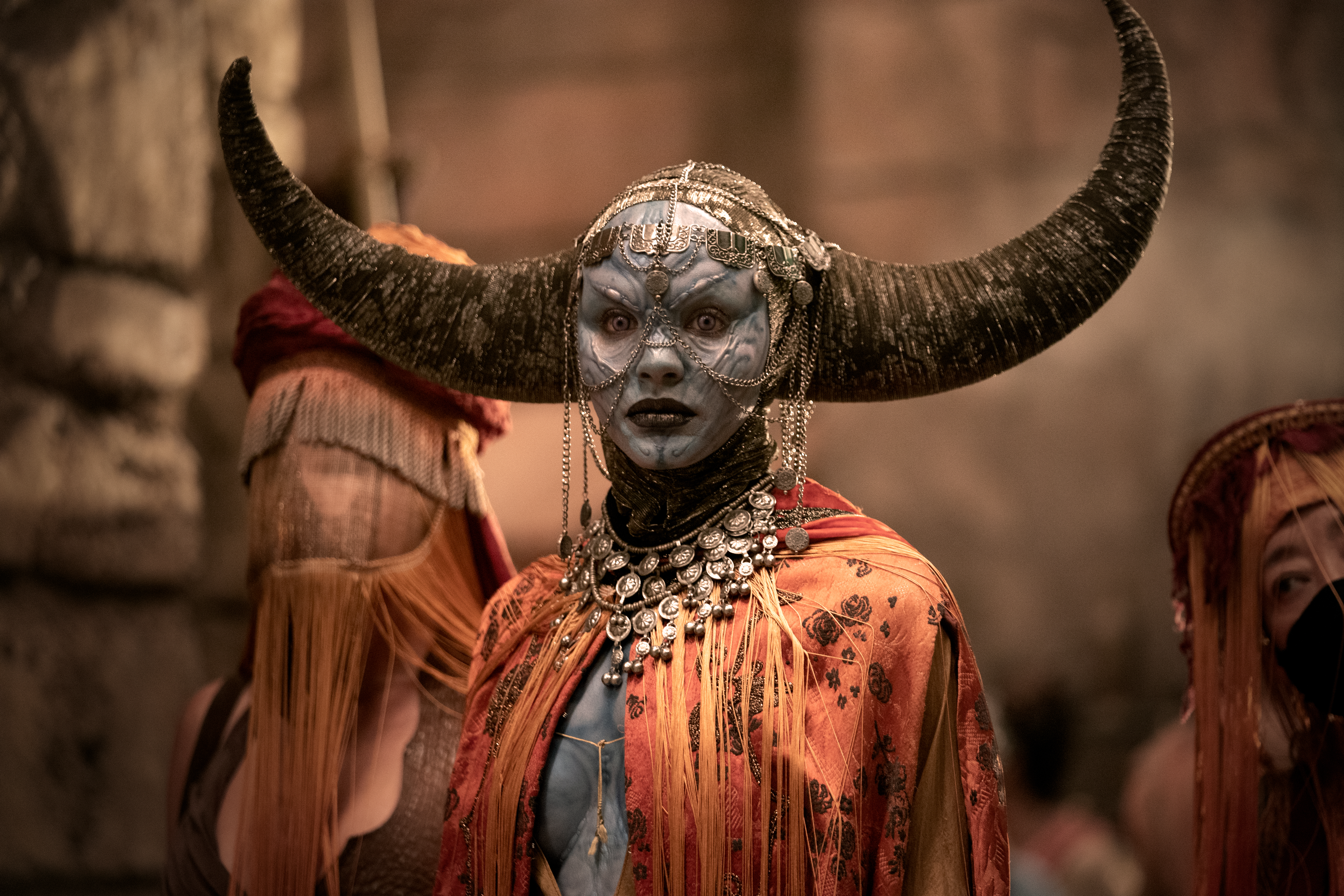 A blue-grey-skinned woman with vast, spreading horns like an ox and her head and shoulders covered in small, fine chains and other jewelry stares directly into the camera in a promo image for Zack Snyder’s Rebel Moon