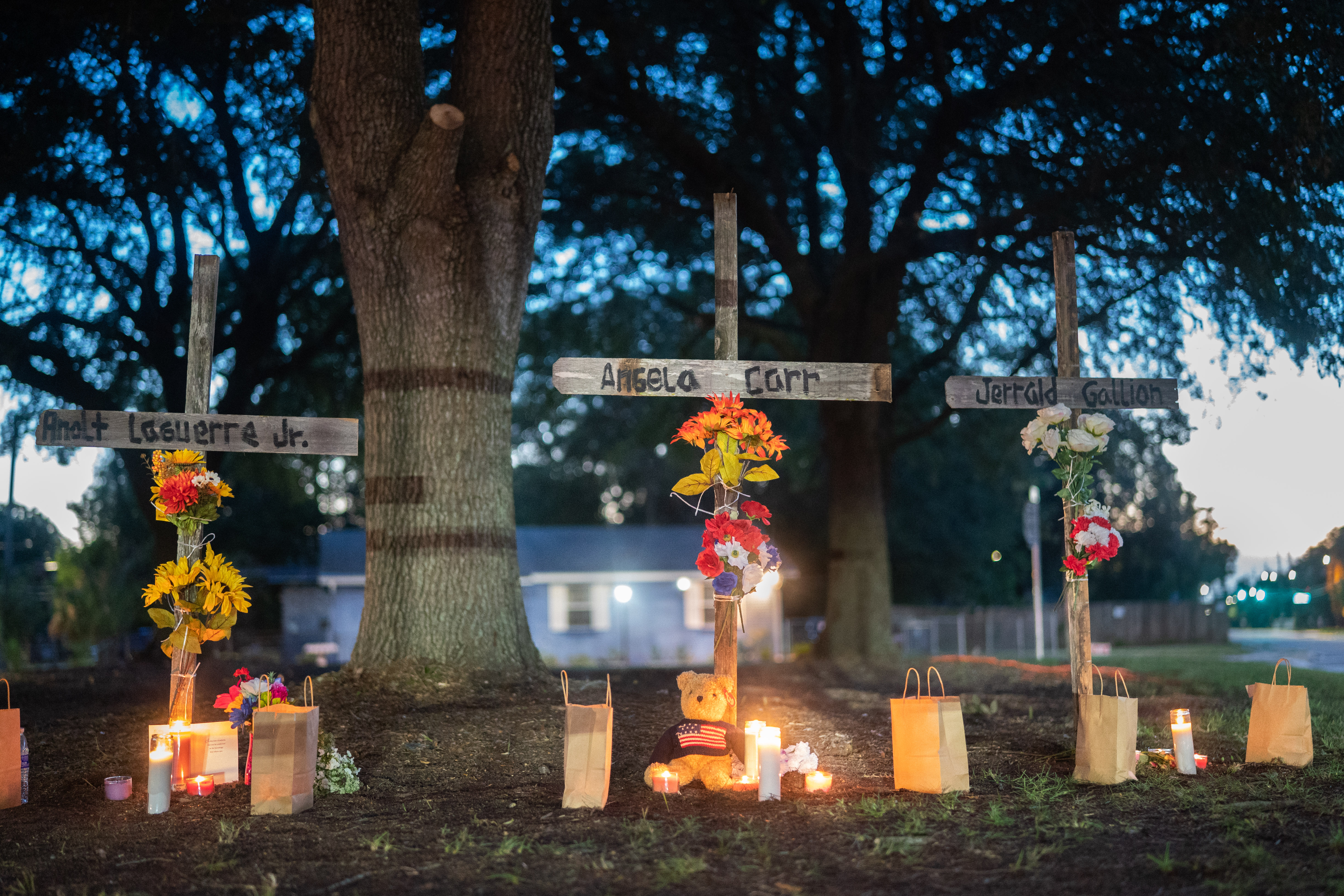 Three white crosses with names stand among tall trees in the yard of a house. Lit candles and luminaria surround them on the ground, with other mementos.