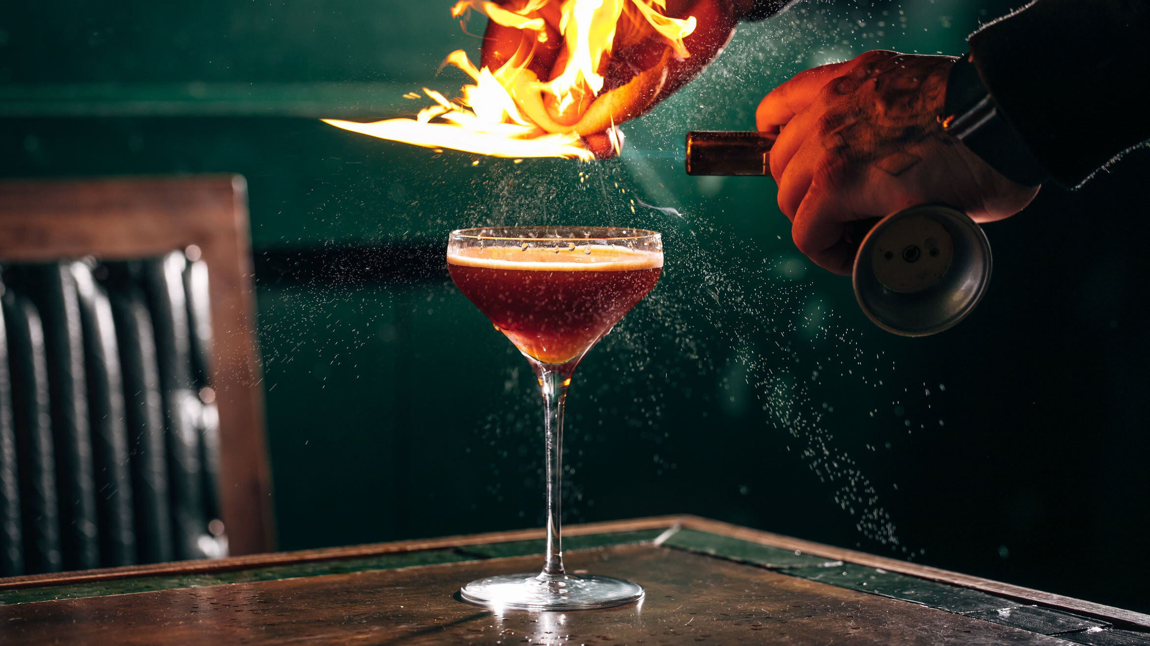 A hand squeezes a flaming citrus wedge into a cocktail glass