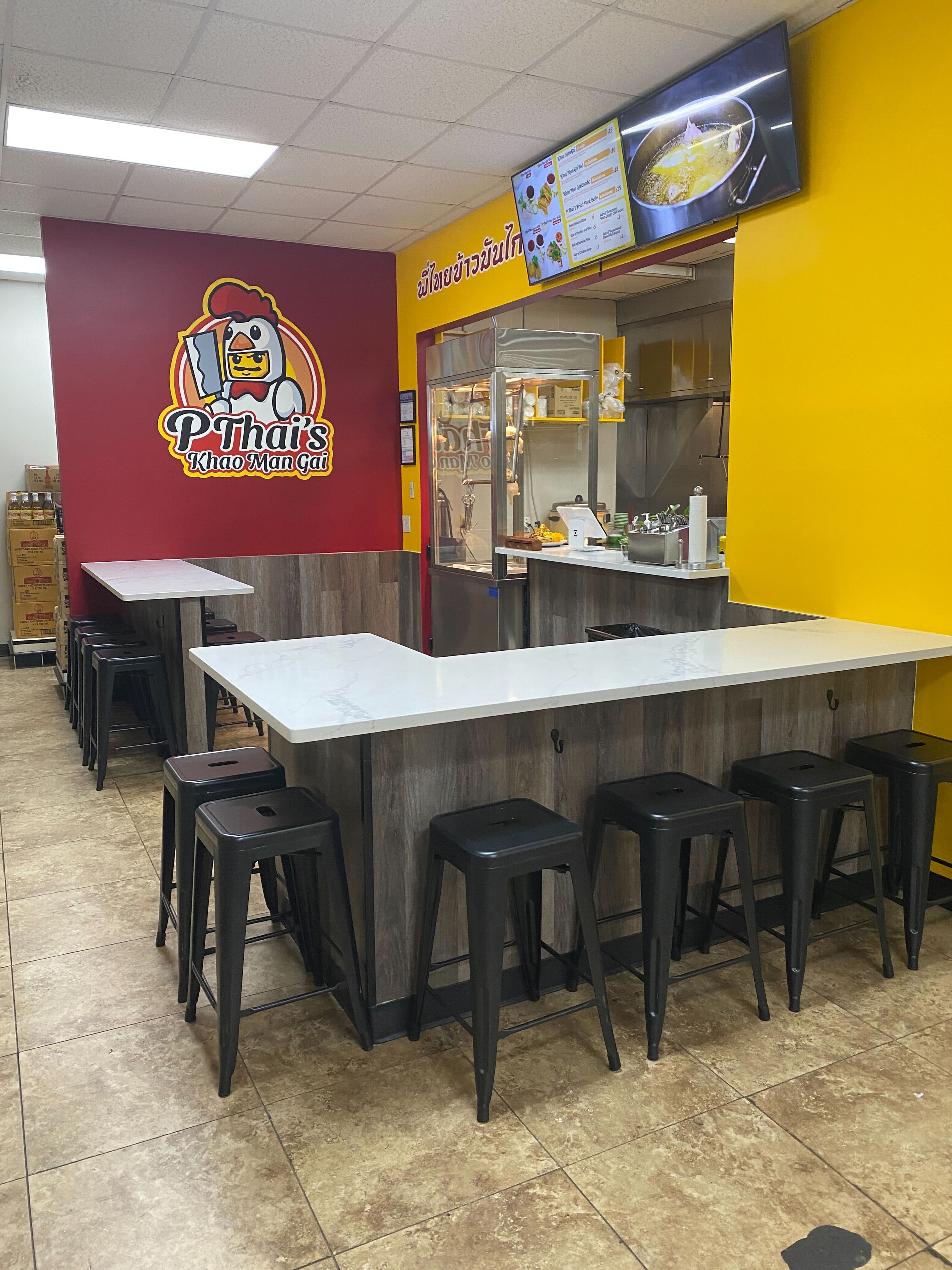A restaurant food stall counter with yellow and red walls, a chicken logo on the red wall, and bar stools.