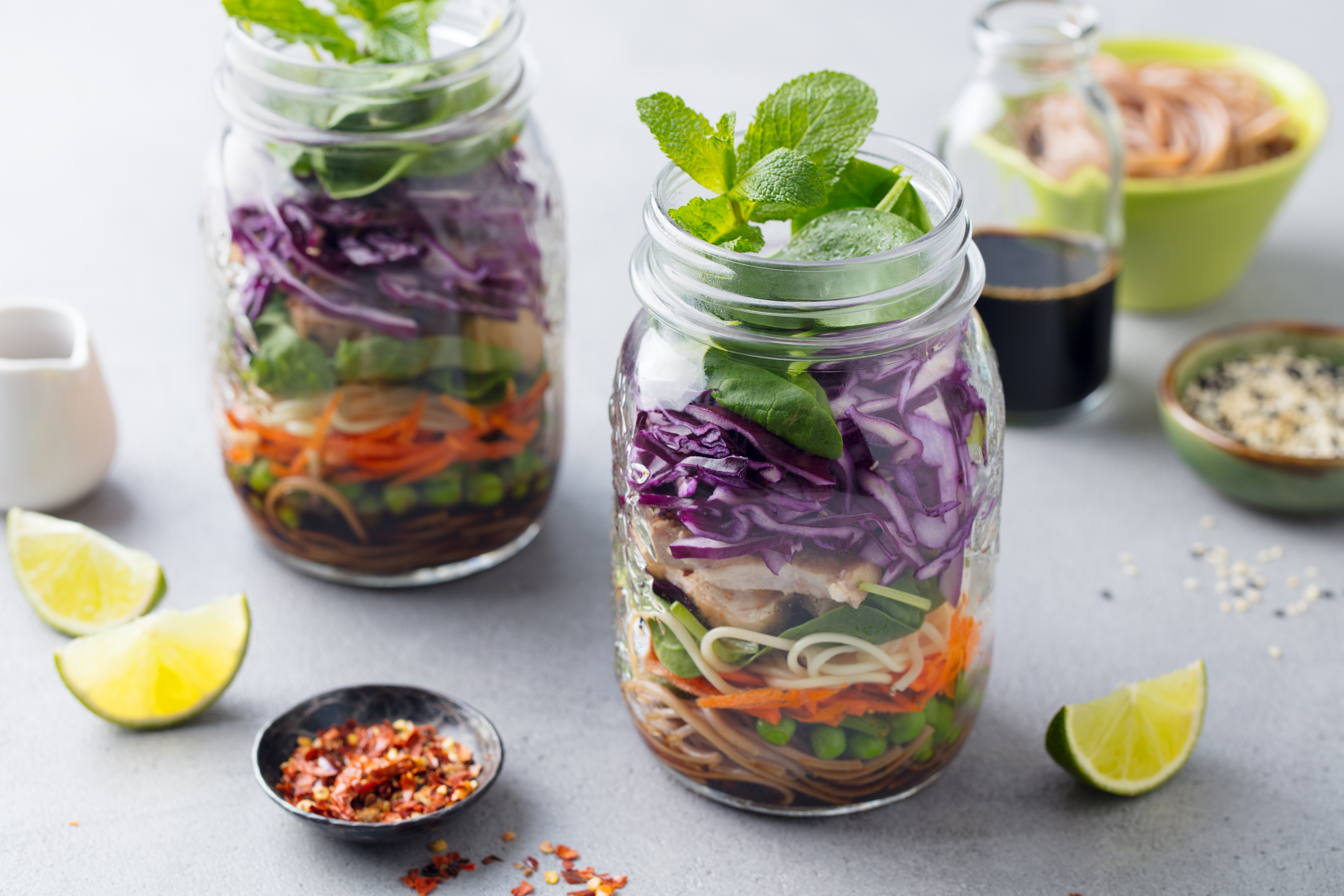 A pair of glass jars filled with noodles, vegetables, and fresh herbs.