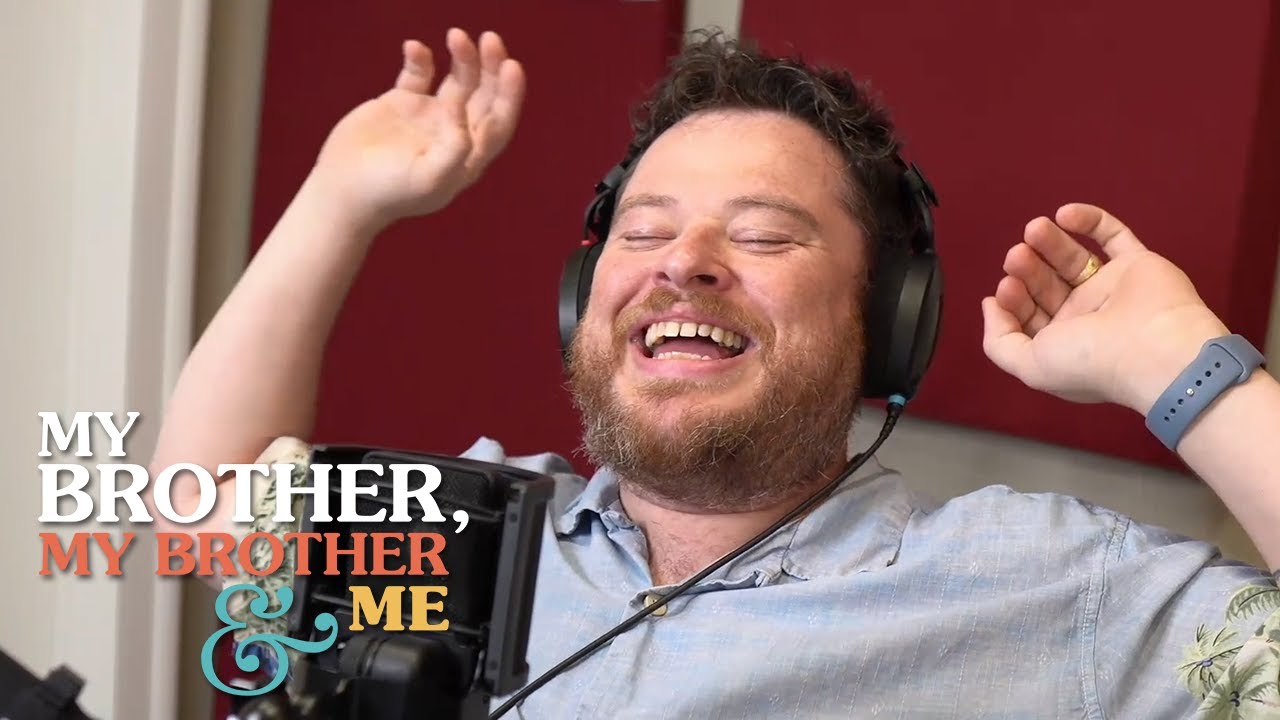 A photograph of Justin McElroy, leaning back laughing, with his hands thrown up near his head. He is wearing headphones and is seated behind a microphone. The MBMBaM logo is superimposed in the lower left corner.