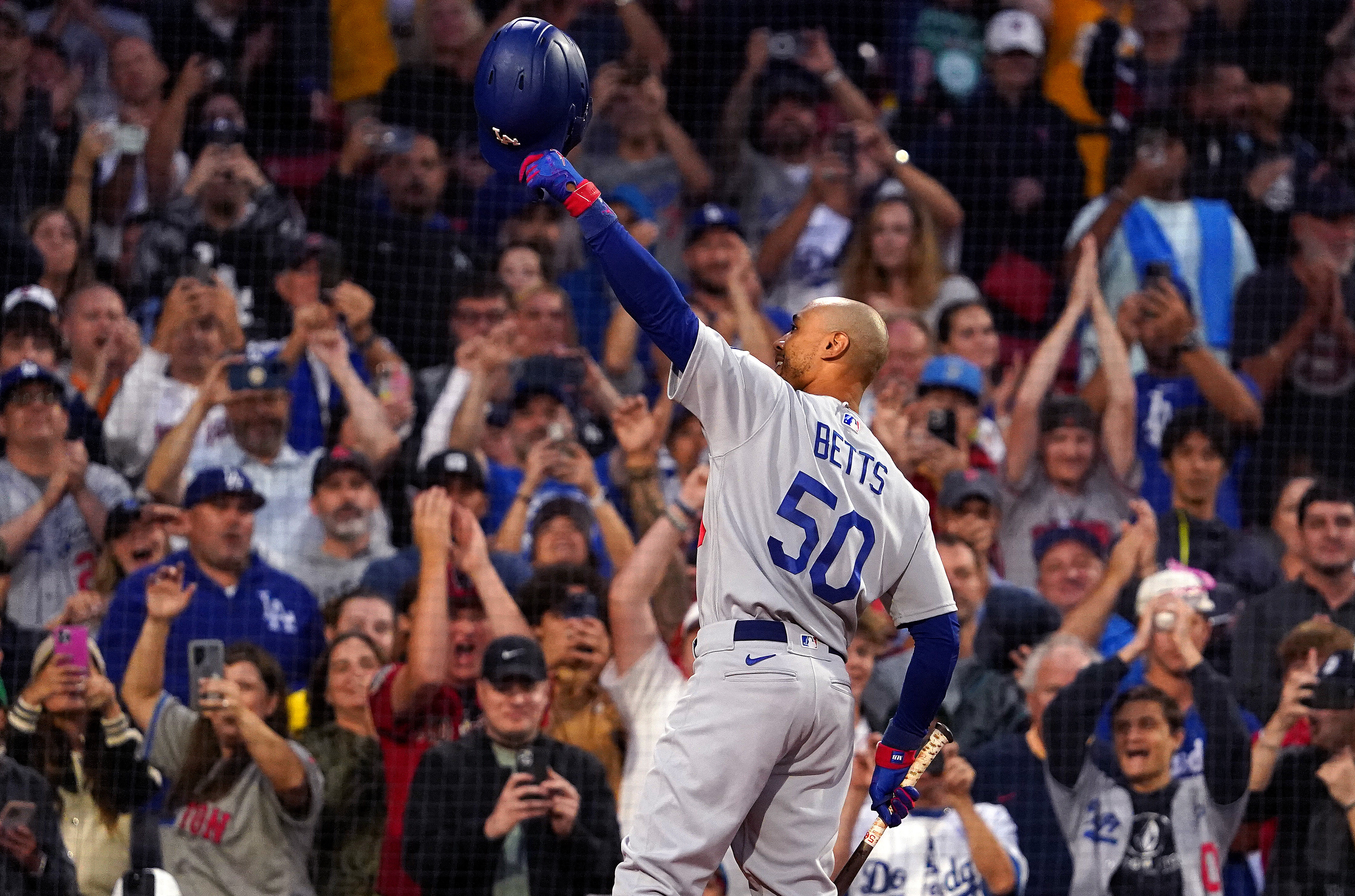Mookie Betts, wearing Dodger grays, waves his helmet to a cheering crowd at a baseball stadium.