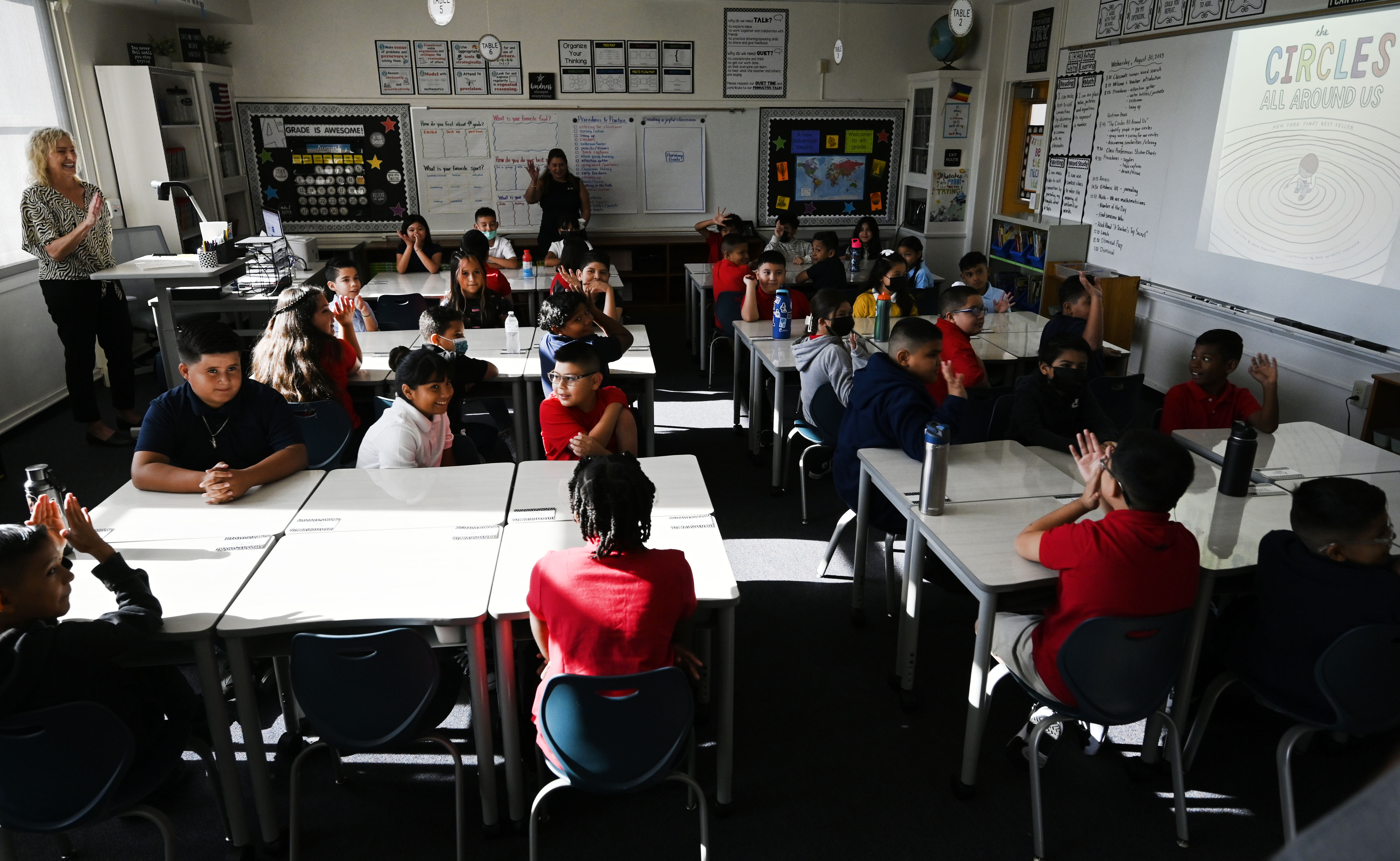 Young students sit at white desks in a classroom while a teacher gives instructions from one side of the room.