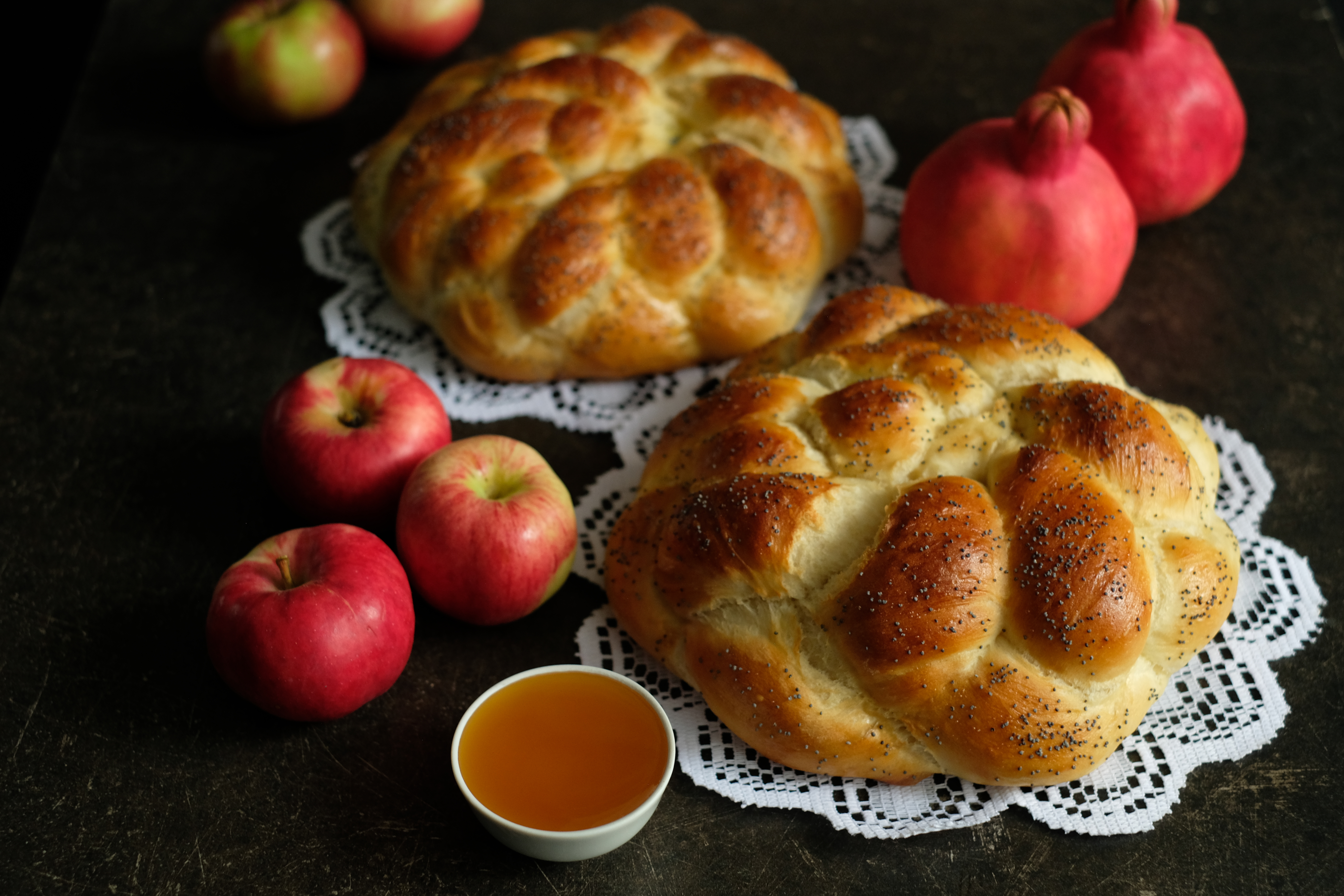 Two crown challah next to apples and a small dish of honey on a black background. 