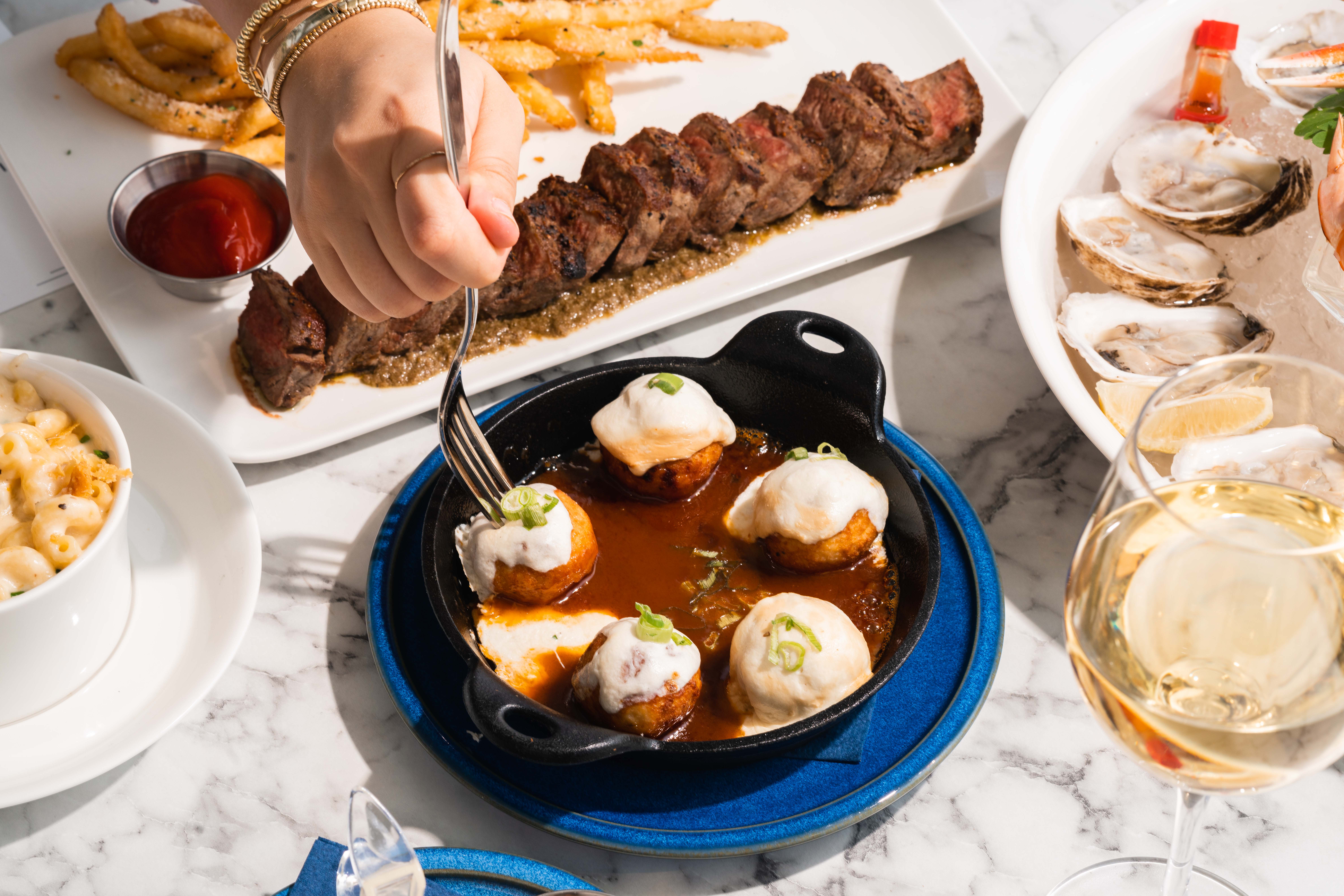 A marbled table with various plates and dishes filled with various food items including oysters, steak and fries, and someone grabbing a meatball from a plate using a fork. 