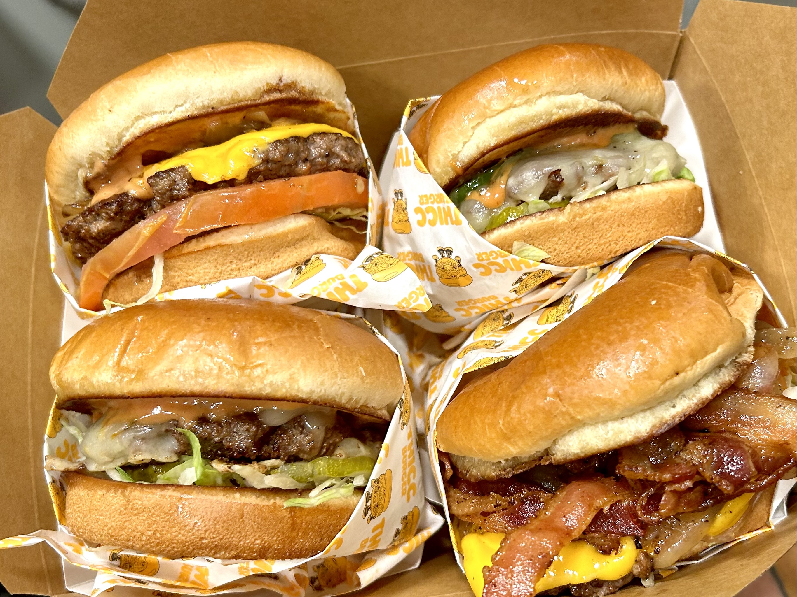 An overhead, slightly tilted shot of burgers wrapped in yellow and white paper at a new stand, with bacon and other toppings.
