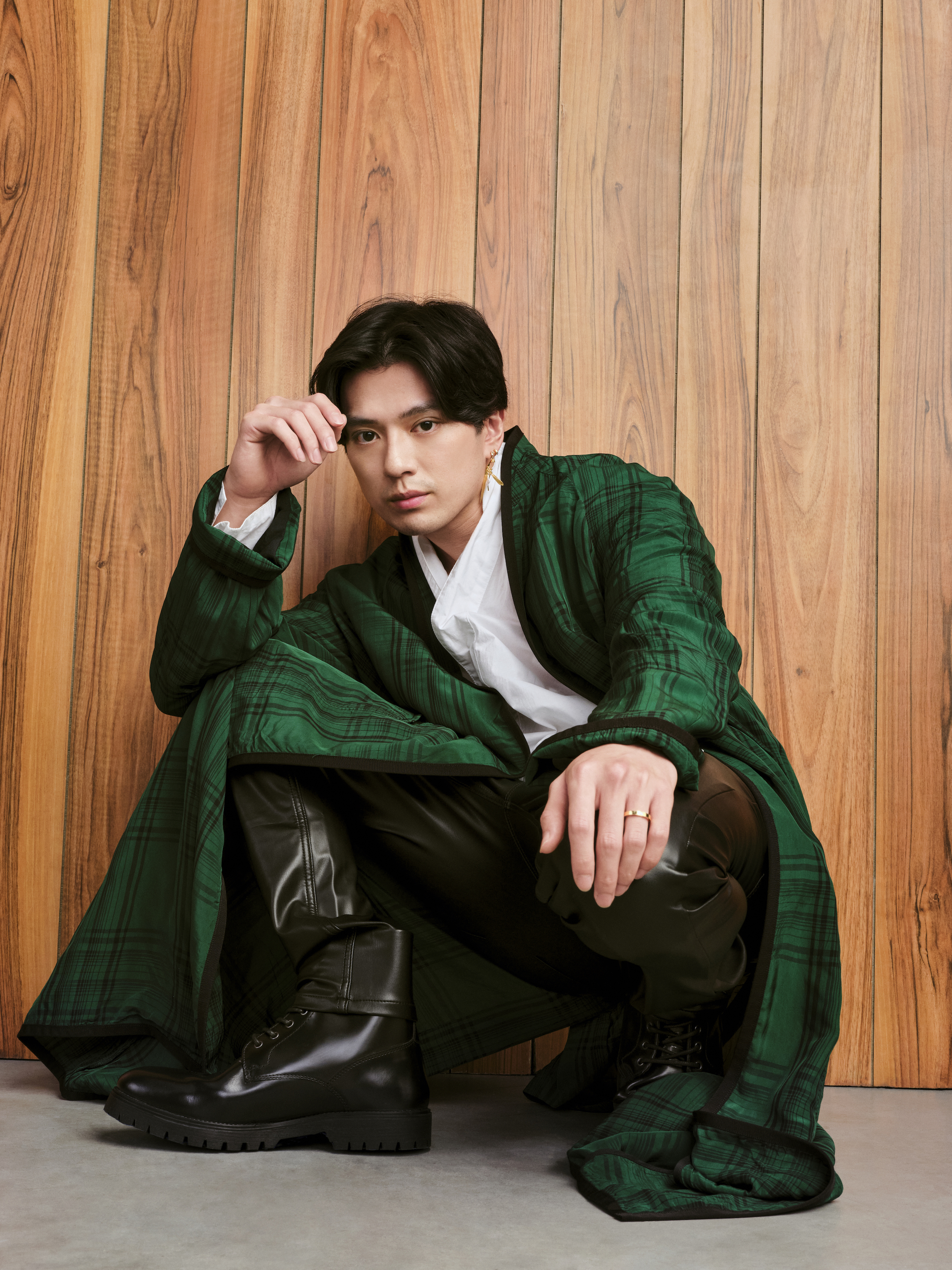 Mackenyu in a green coat giving a fashion cover pose while leaning against a wood wall in a One Piece photo shoot