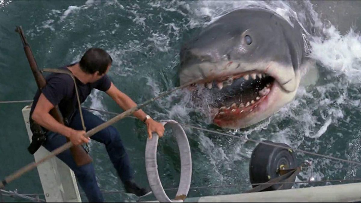 A man with a spear stands on the side of a boat tipping into the sea while staring into the eyes of Great White Shark barring its teeth in Jaws.