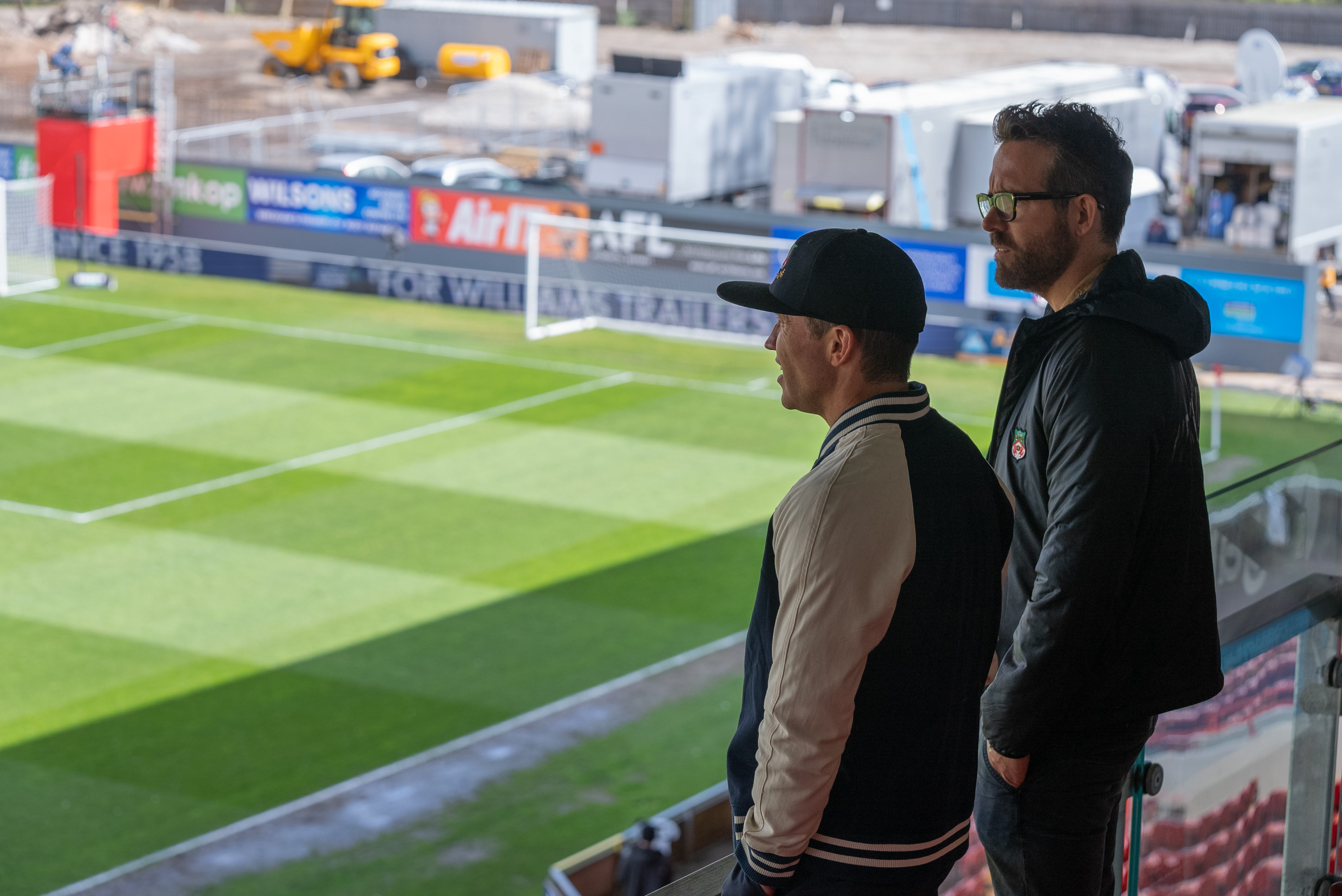 Rob McElhenney and Ryan Reynolds stand in the seats of a soccer stadium looking out over the pitch.