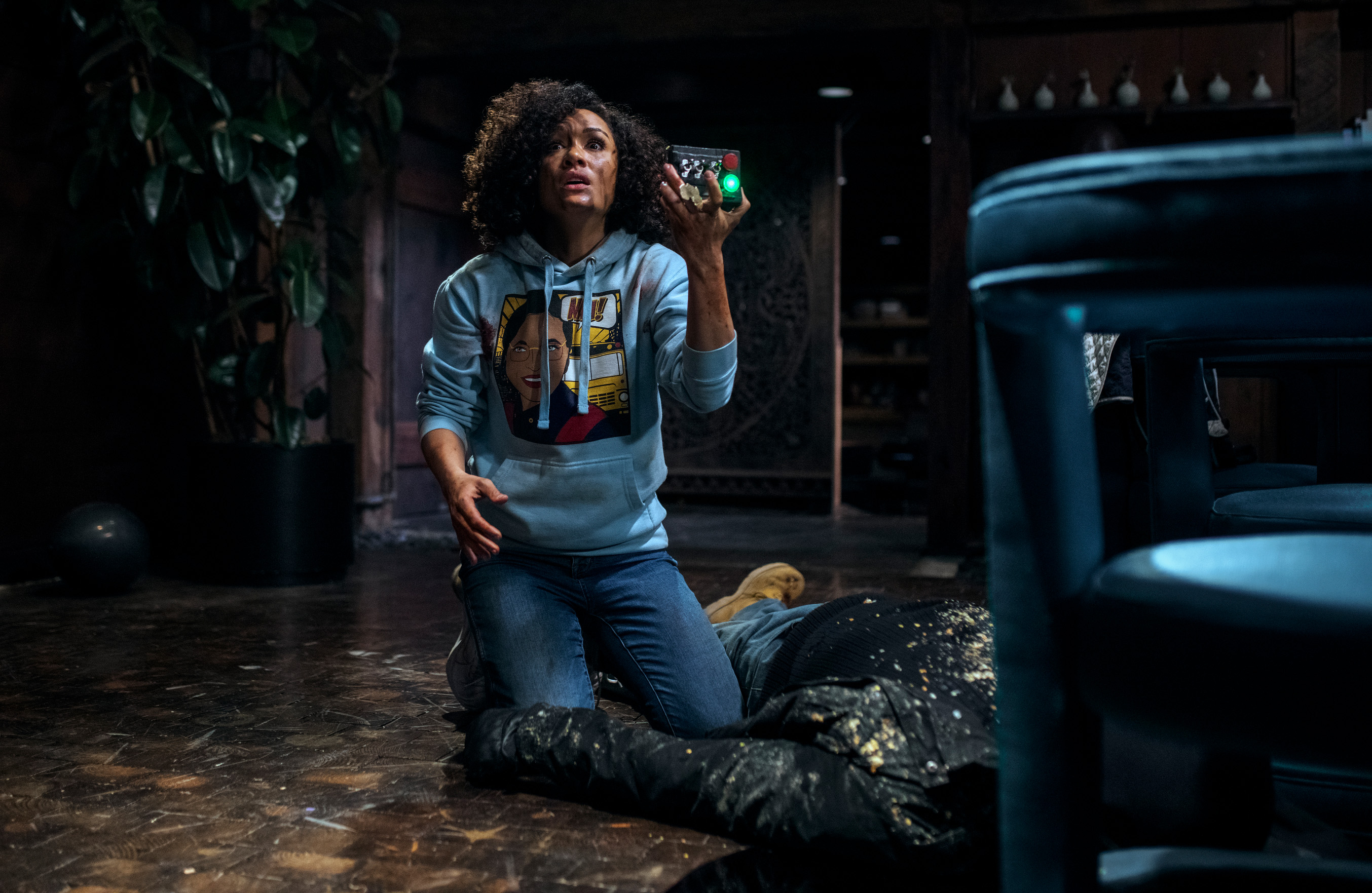 A woman in blue sweatshirt and jeans and dirt on her face stands over a body in an old cabin while holding a box with switches and a green light in The Blackening