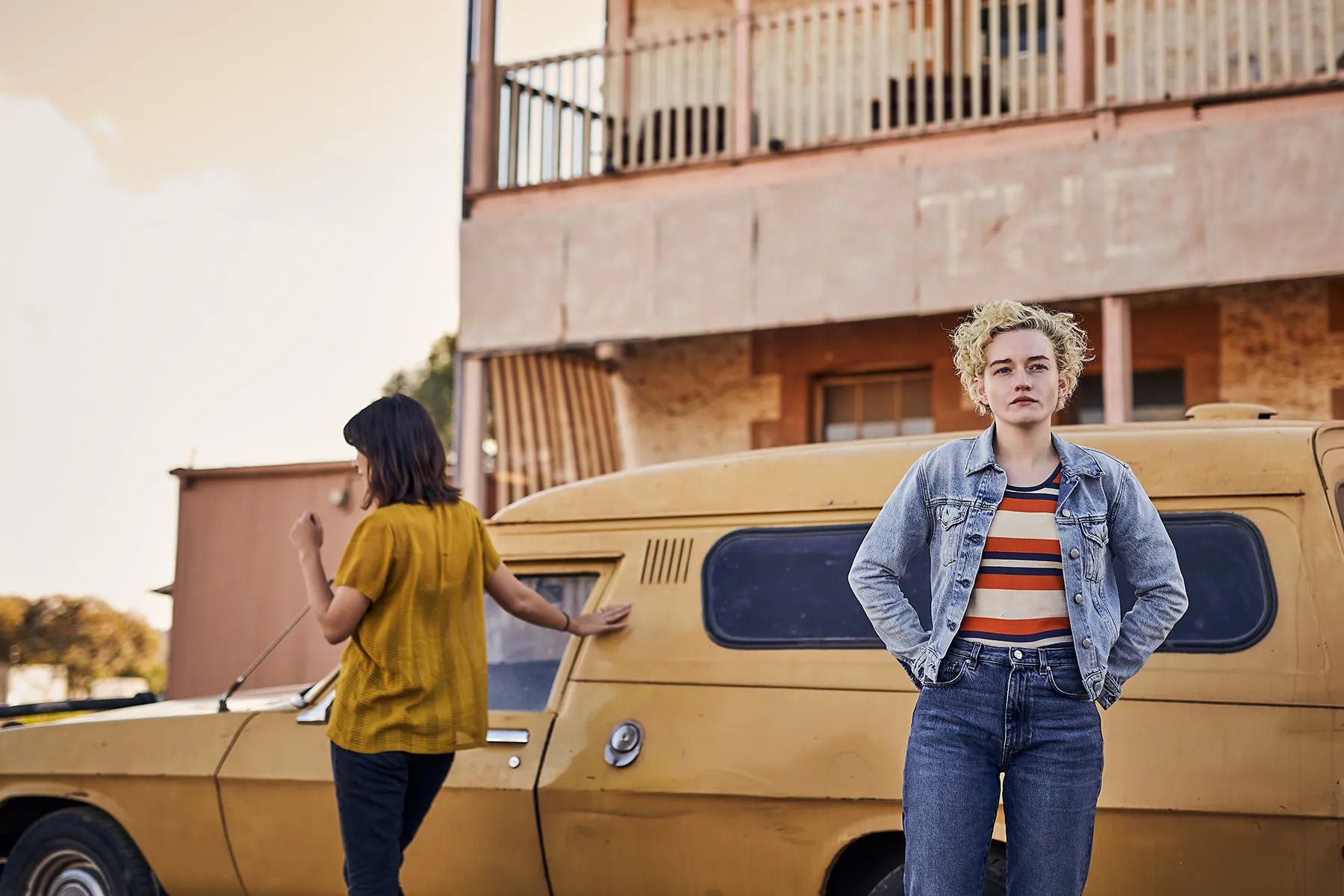 Two yong women stand in front of a yellow car parked in front of a rustic hotel.