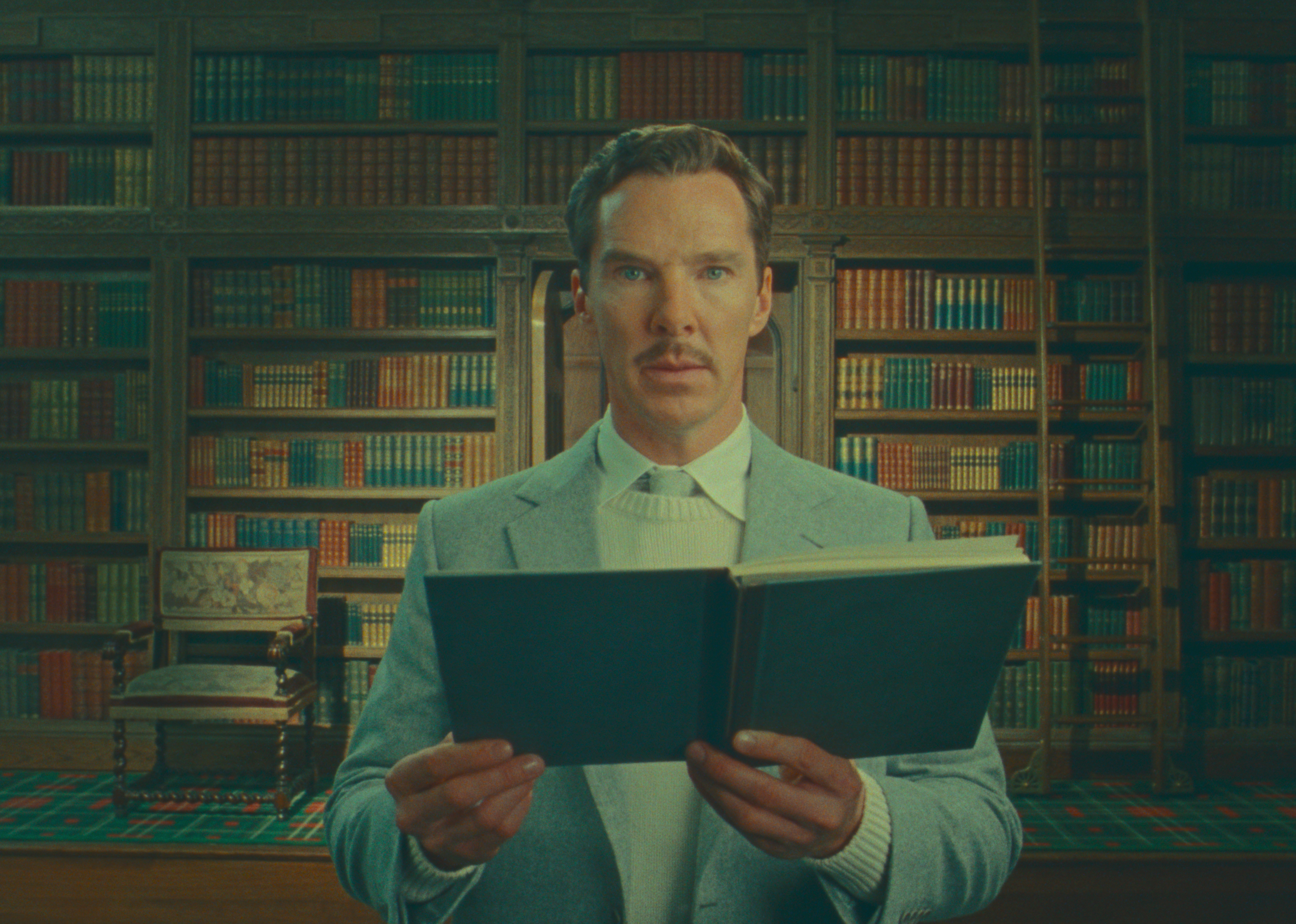 Benedict Cumberbatch, standing in a room lined ceiling to floor with books, stares directly into the camera while holding up another book in Wes Anderson’s Netflix film The Wonderful Story of Henry Sugar