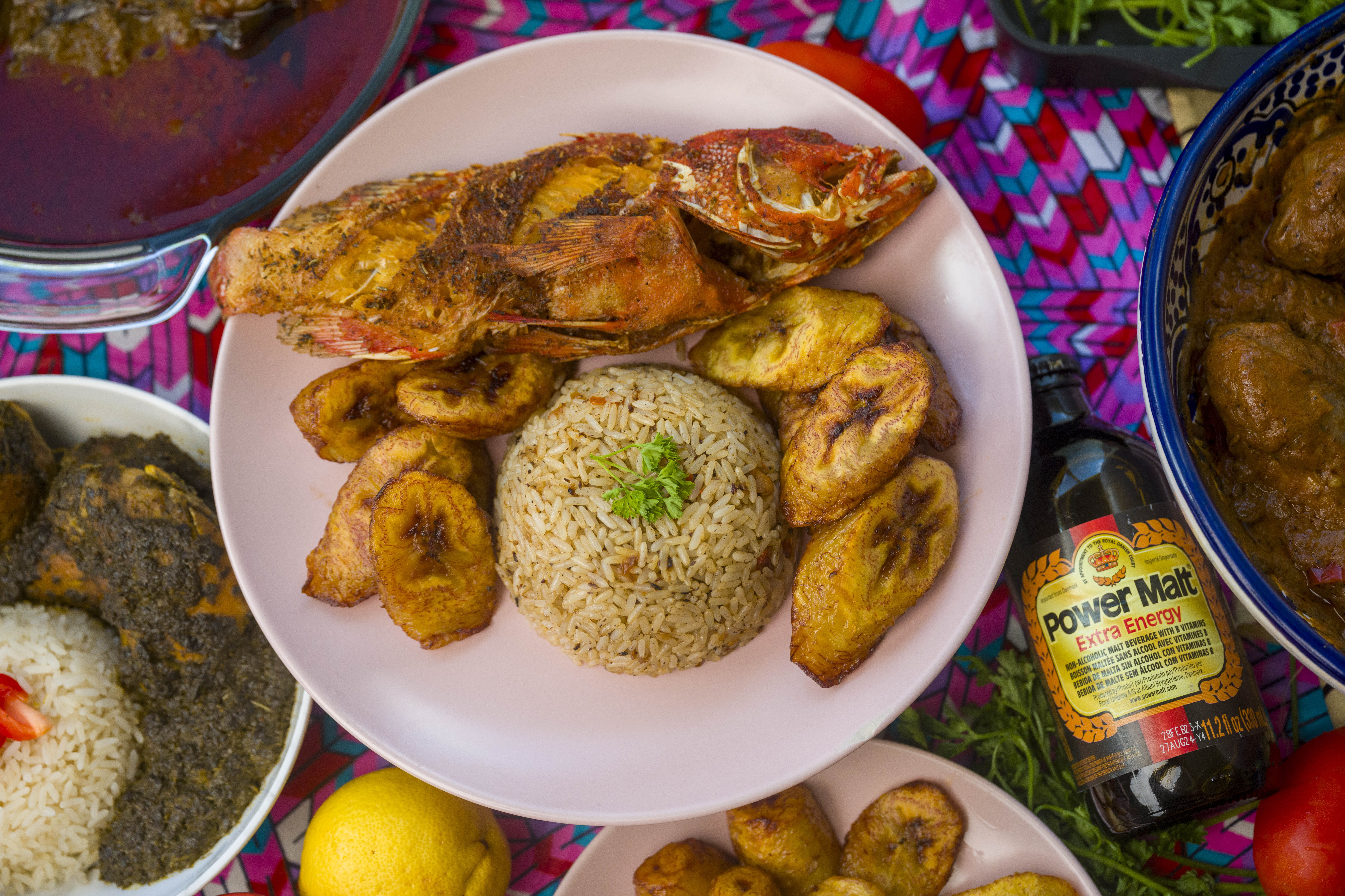 A whole fried fish with plantains and rice on the side on a round plate surrounded by other dishes set on a multi-colored table cloth.
