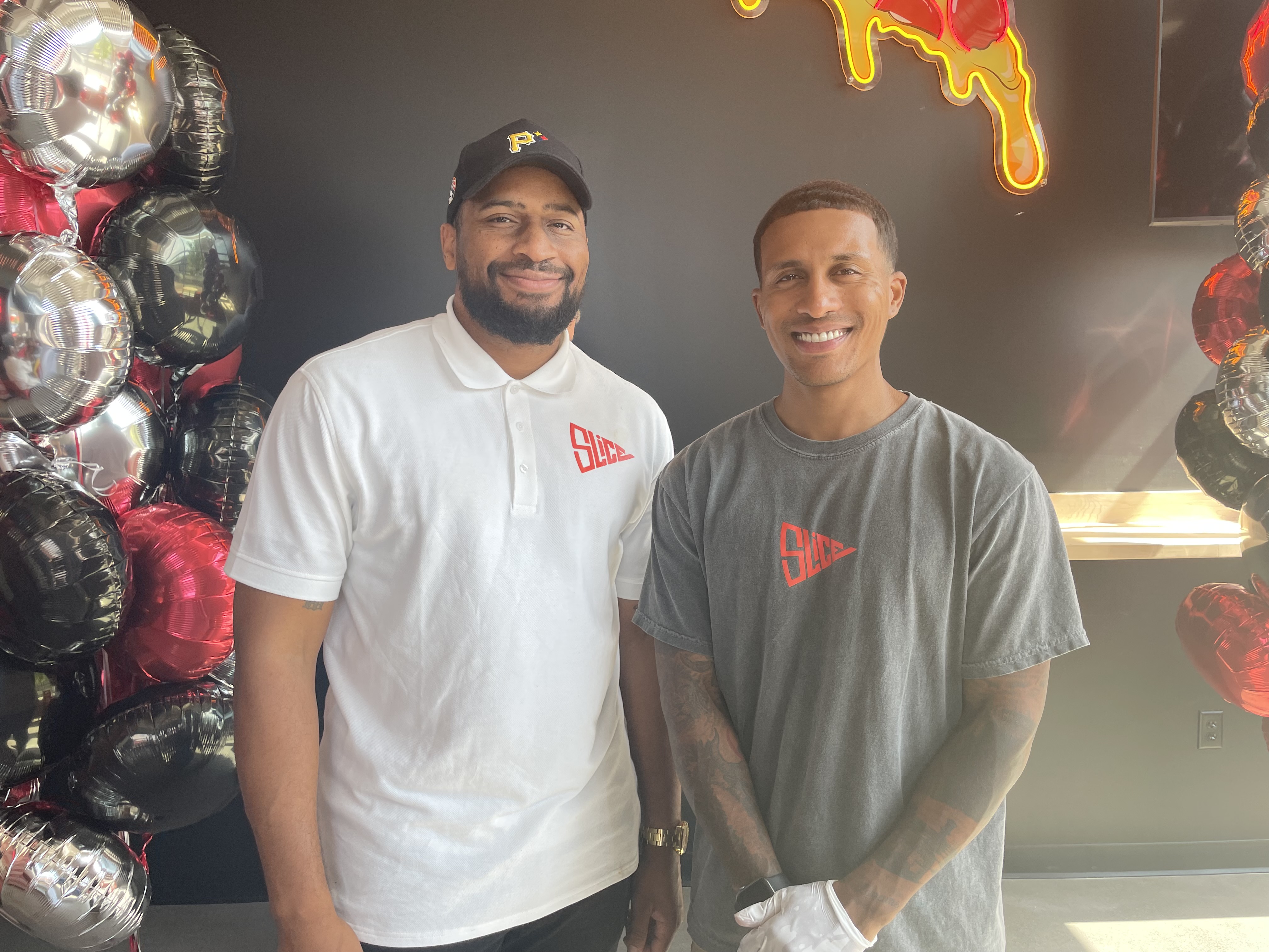 Owners Hosie Thurmond and Adam Kado wearing shirts with a small orange label that says “Slice” against a black wall with red, black, and silver balloons visible on either side. 
