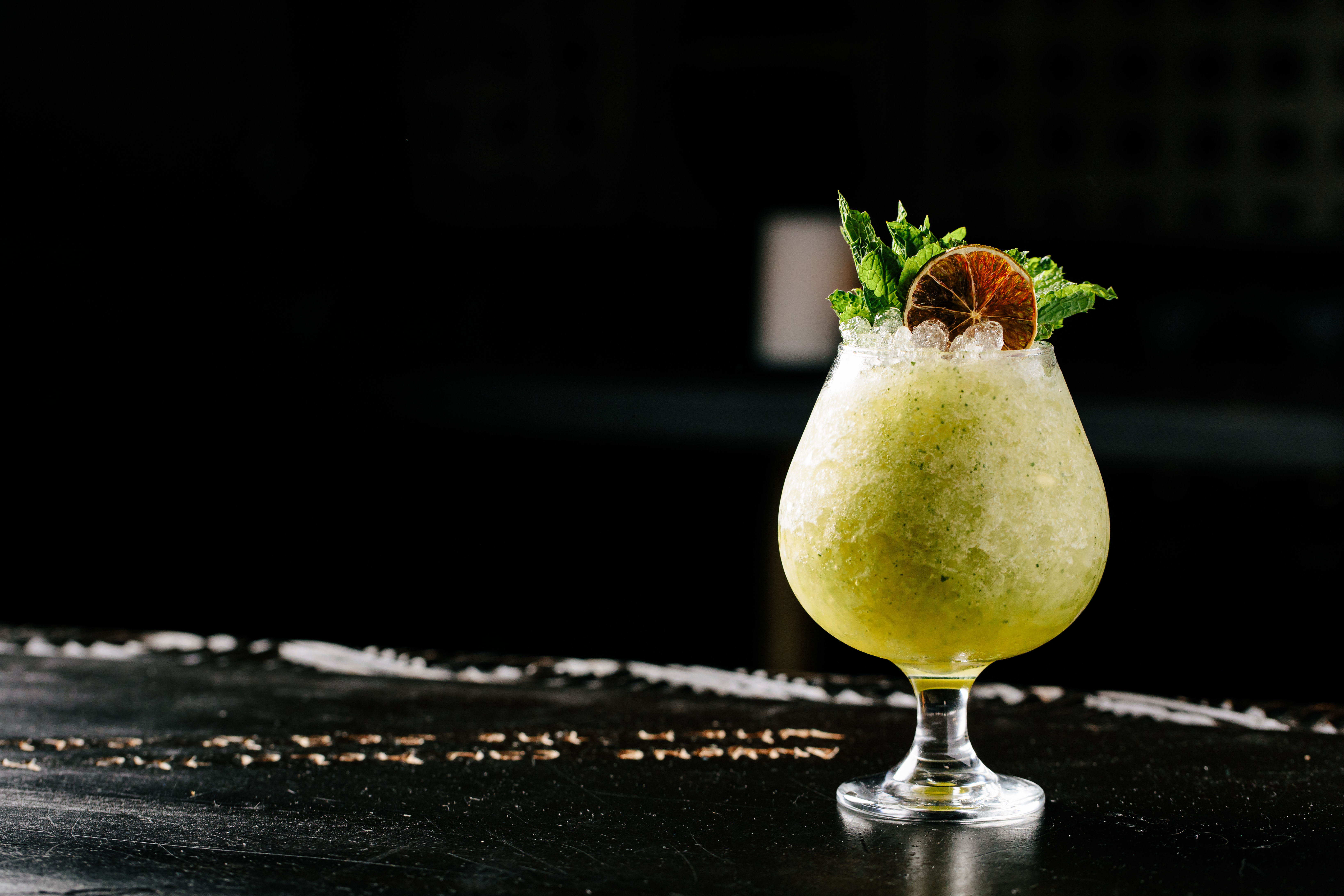 A greenish cocktail with lots of ice and green garnishes in a clear goblet glass on a black surface.